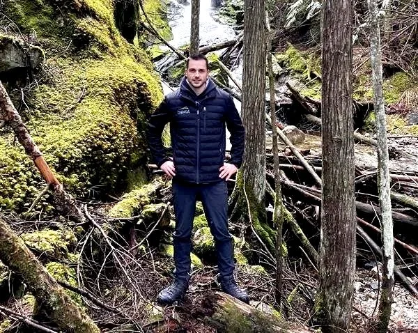 A man in a dark jacket and pants standing in a dense forest with moss-covered trees and fallen logs.