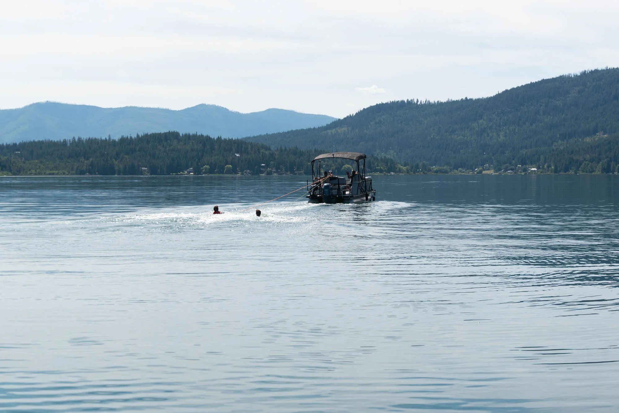 A motorboat on a calm lake with two people in the water, surrounded by forested mountains under a cloudy sky.
