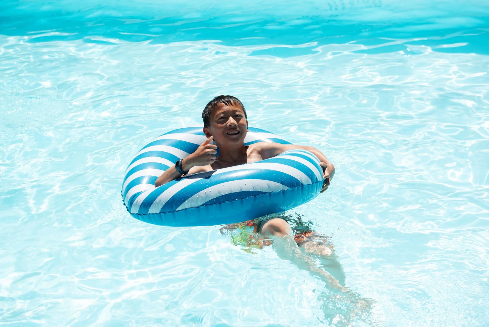 A young man in a swimming pool floating on a blue and white striped inflatable ring, smiling, with water and sunlight reflections surrounding him.