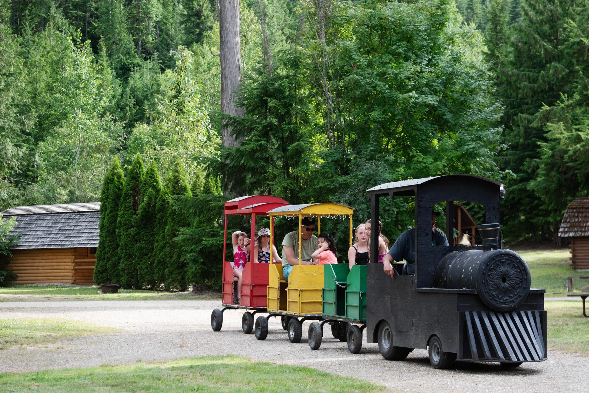 Children riding a small train through a green, forested area with trees and cottages.