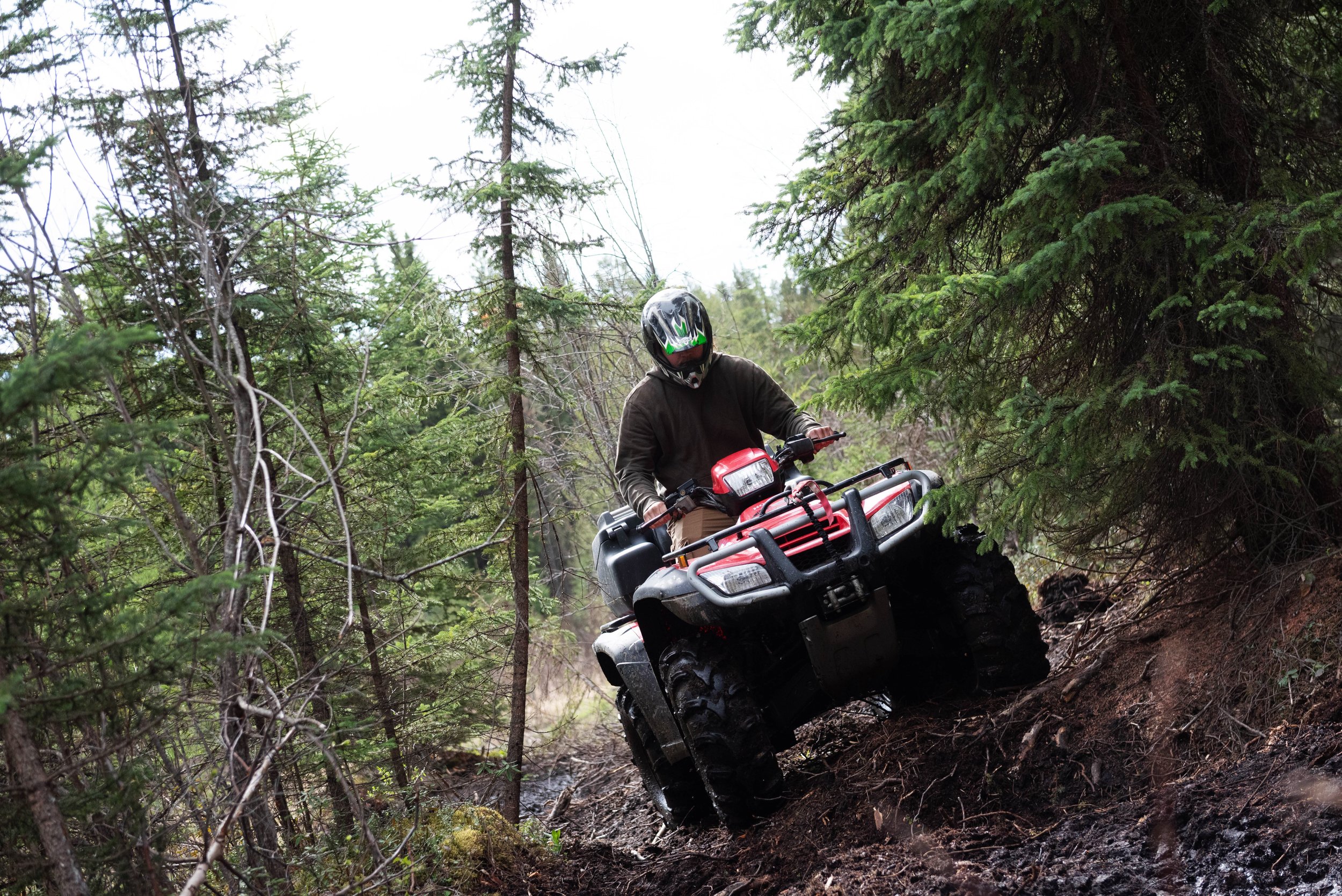 A person riding an all-terrain vehicle (ATV) through a forest trail among green trees, wearing a helmet and dark clothing.