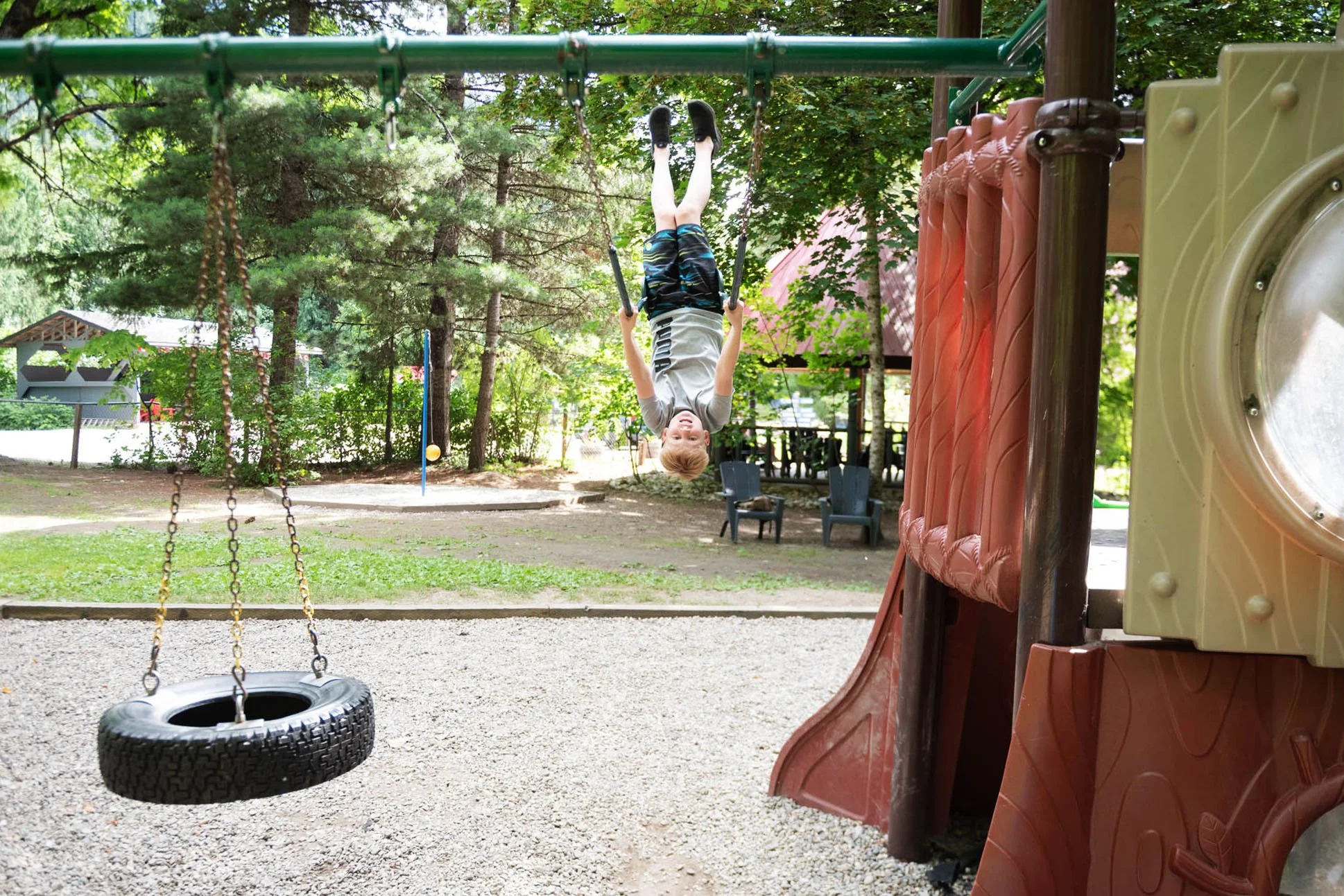 A young boy hanging upside down on a playground, with a tire swing visible in the foreground. The playground is outdoors, surrounded by trees and greenery.
