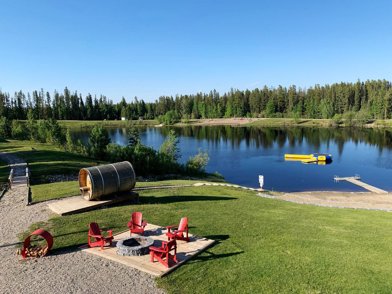 Scenic view of a lake with a dock, yellow paddle boats, a grassy area with red chairs around a fire pit, and a hillside with trees under a clear blue sky.