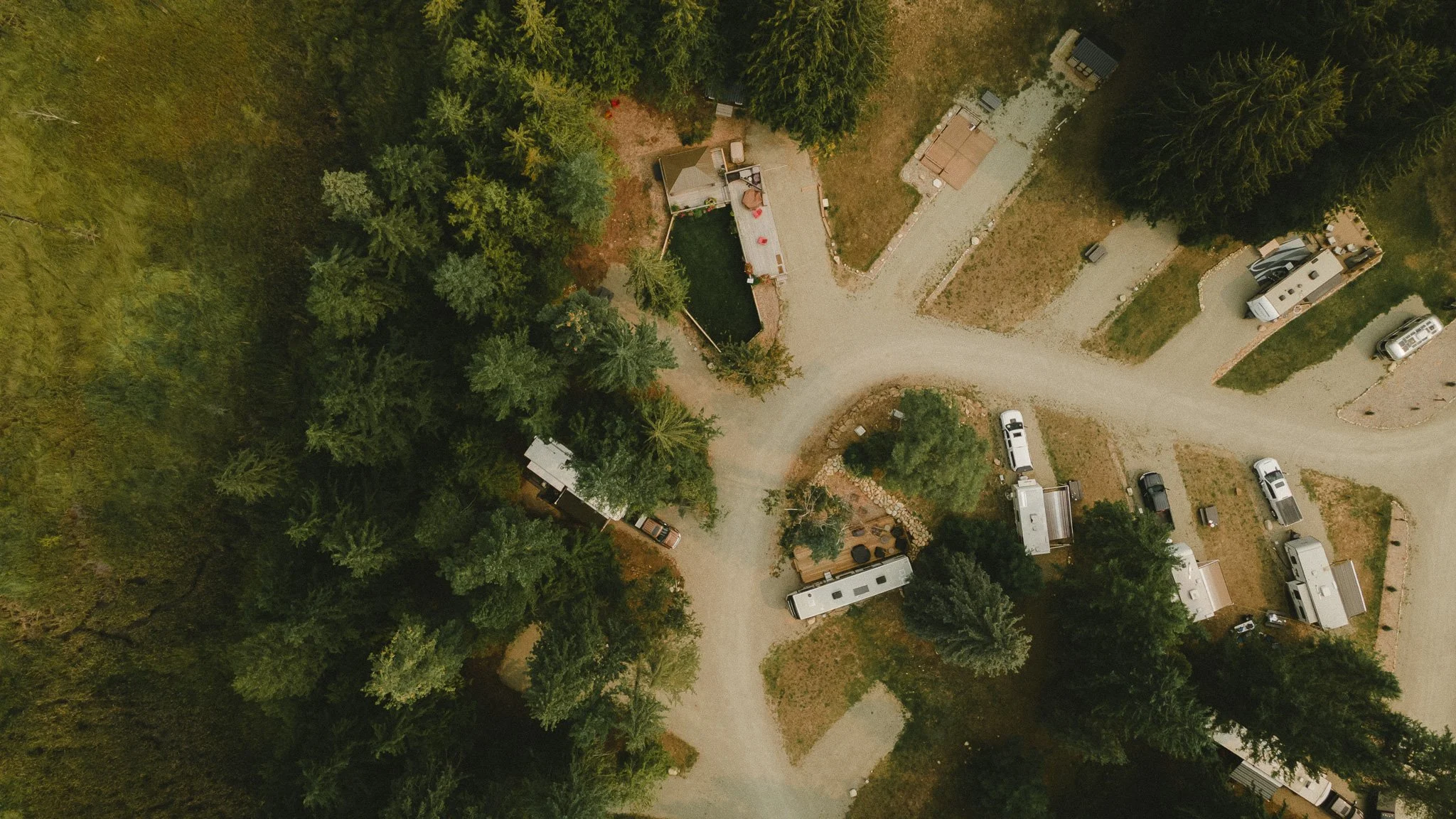 An aerial view of a wooded area with several parked RVs and trailers along a dirt road, some small structures, and a house with a deck.