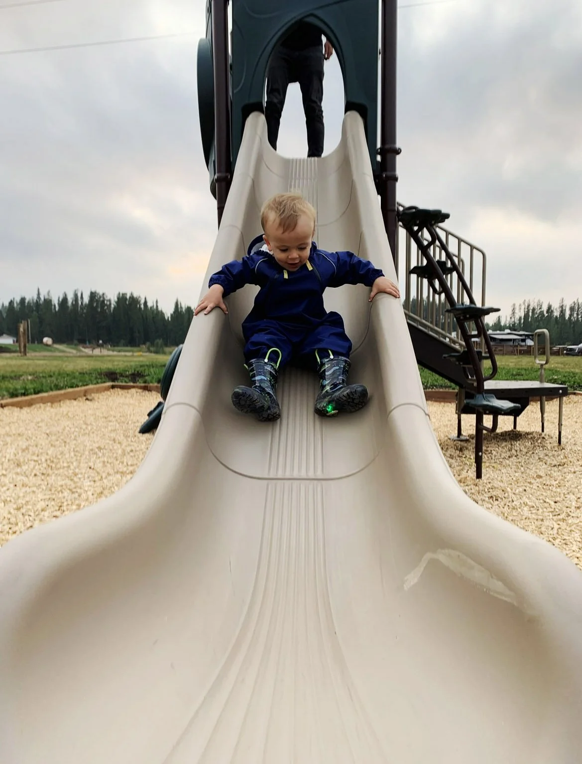 A young child in a blue jacket and rain boots sliding down a playground slide with a person at the top of the slide.