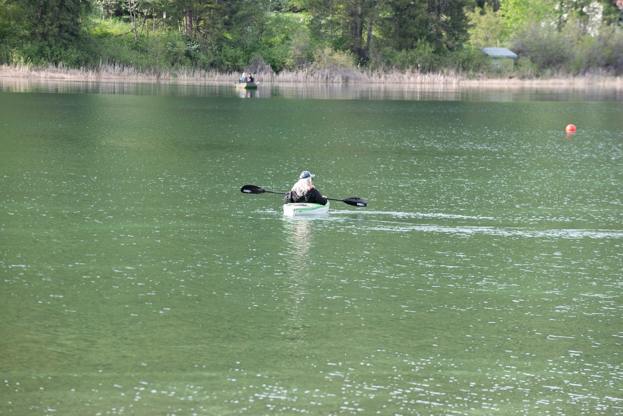 A woman kayaking on a green lake, with a boat carrying two people in the background near the shore of a wooded area.