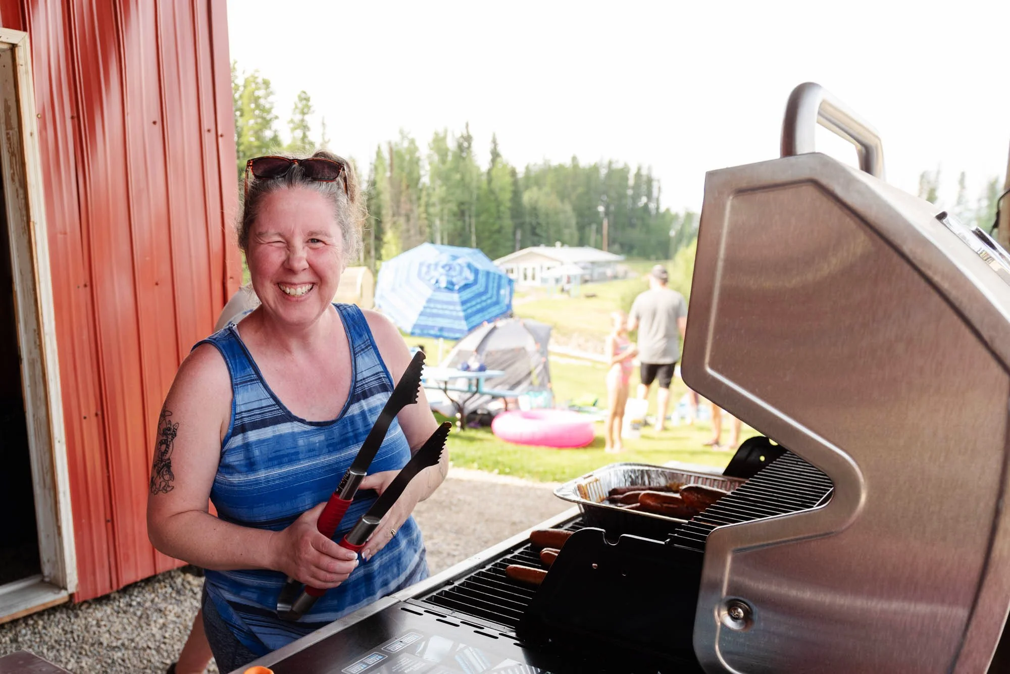 A woman smiling and wearing sunglasses on her head, grilling hot dogs on a barbecue grill at an outdoor gathering.