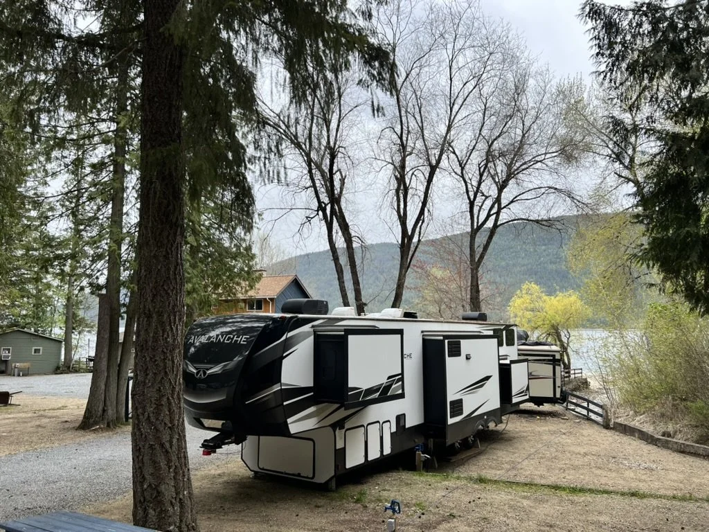 A white travel trailer with black accents parked among trees near a lake, with mountains and a wooden house in the background.