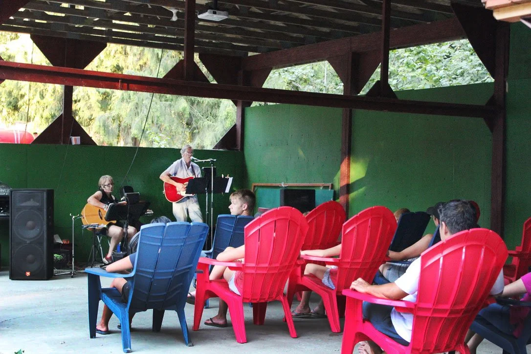 Children sitting on colorful chairs and watching a live outdoor performance by two musicians, one playing a guitar and the other singing with a microphone, under a wooden pavilion.