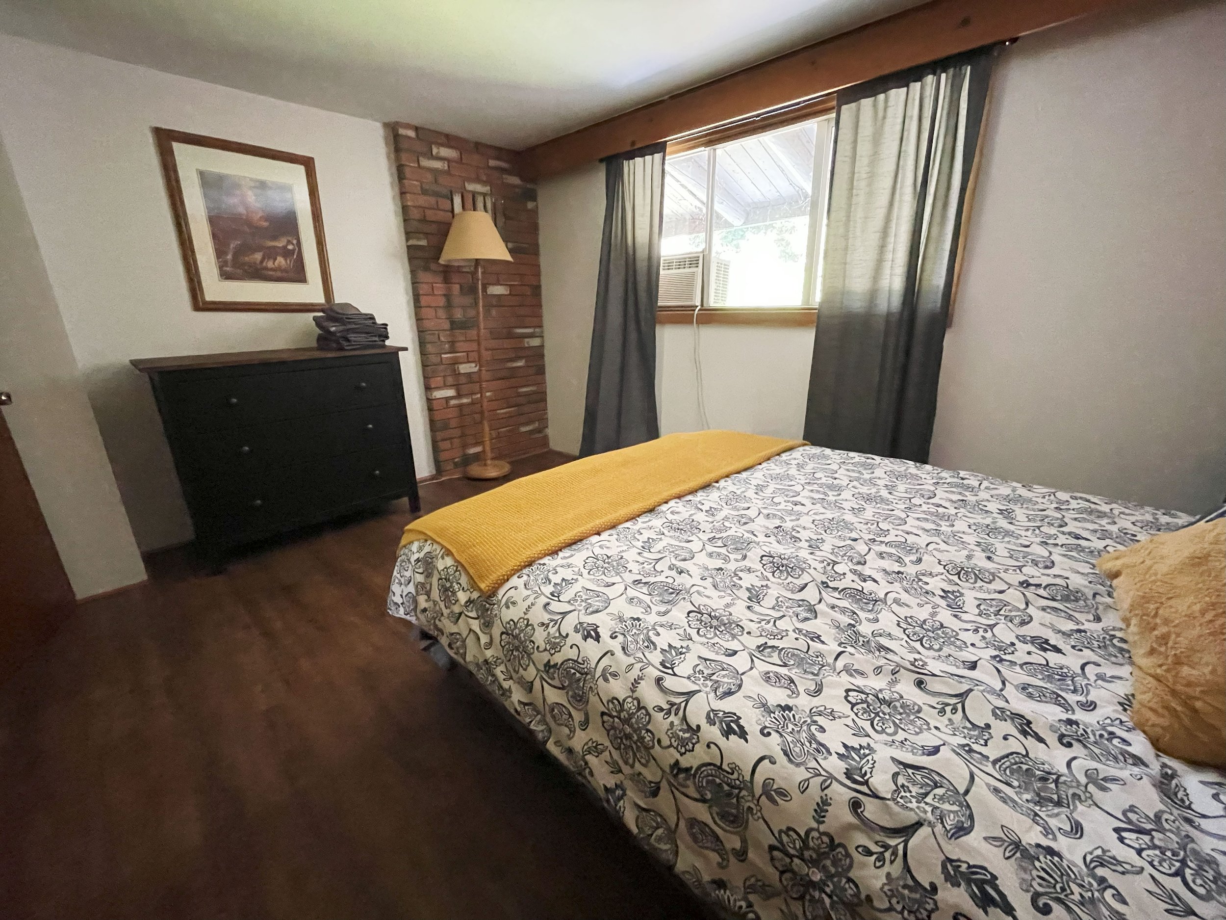 A bedroom with a bed covered in a black and white floral patterned bedspread, a yellow blanket, and a gold pillow. There is a black dresser with folded clothes on top, a framed picture on the wall, a brick corner with a wooden lamp, and a window with