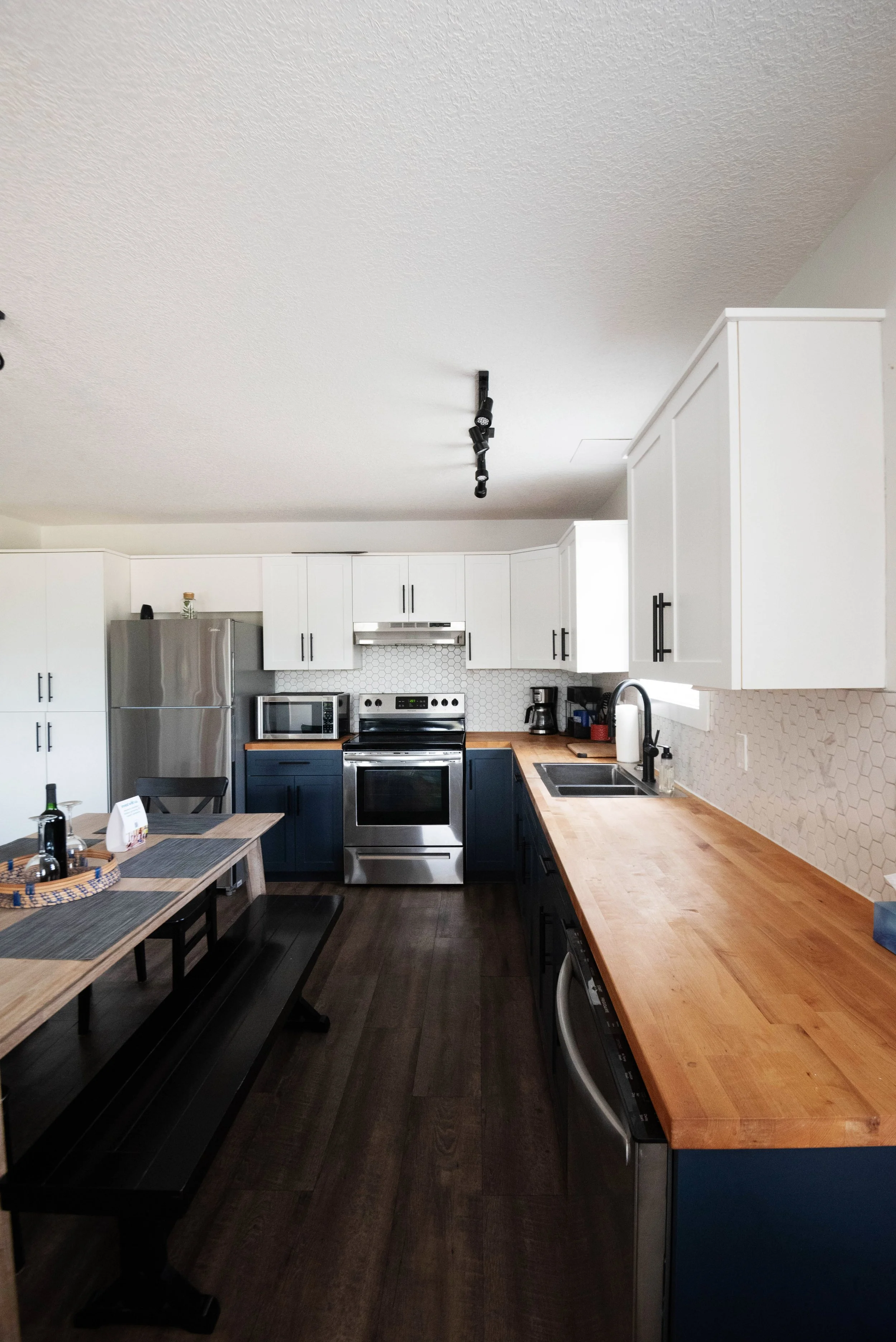 A modern kitchen with white upper cabinets, navy blue lower cabinets, and a wooden countertop. Kitchen appliances include a stainless steel refrigerator, oven, microwave, and coffee maker. A dining table with black benches and a basket with bottles and napkins is also visible.