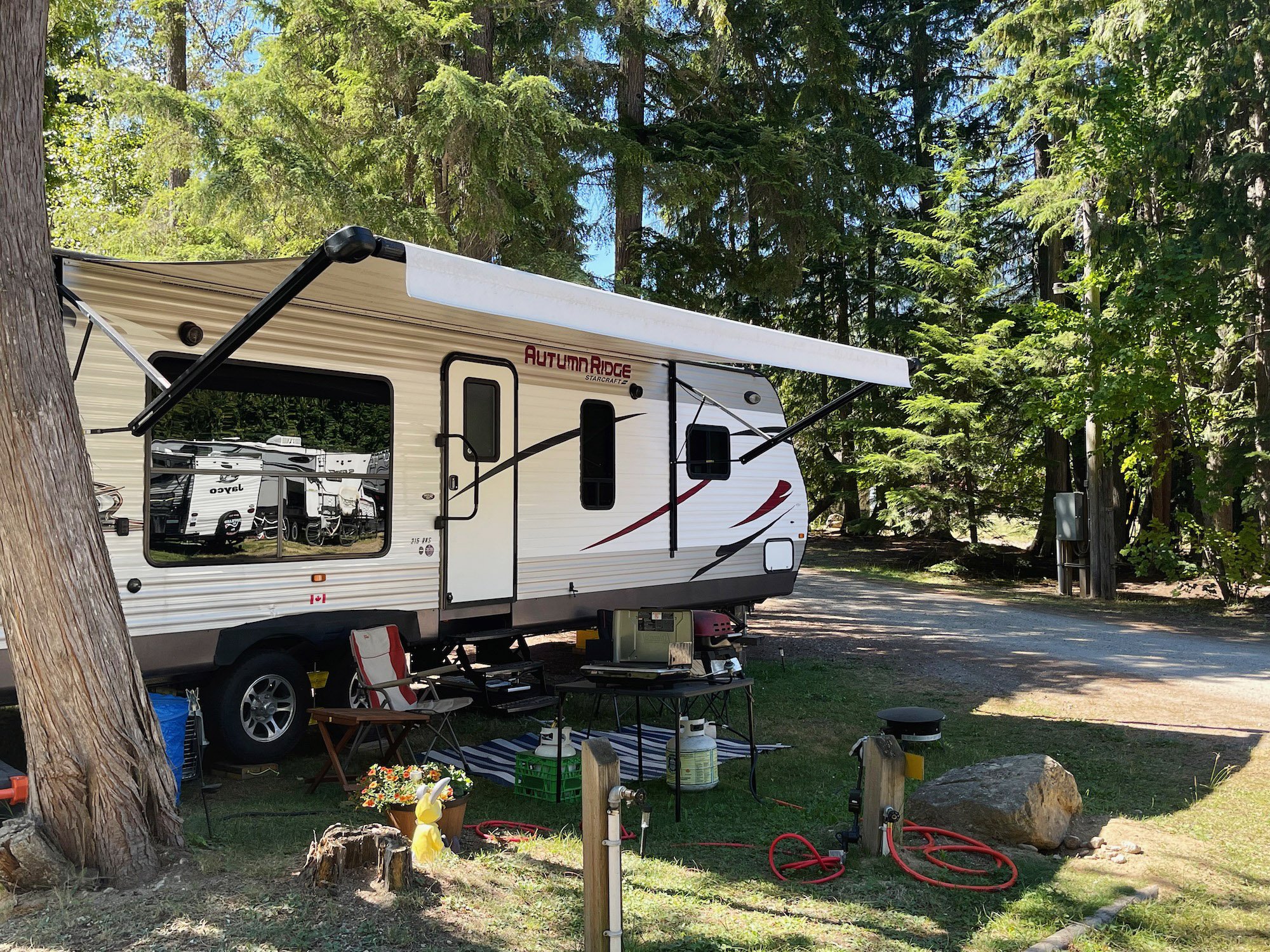 A white Starcraft Autumn Ridge travel trailer parked among tall green pine trees in a wooded campsite. The trailer has a window and an extended awning. The campsite has various outdoor furniture, potted plants, a small table, and camping supplies. Dappled sunlight filters through the trees.
