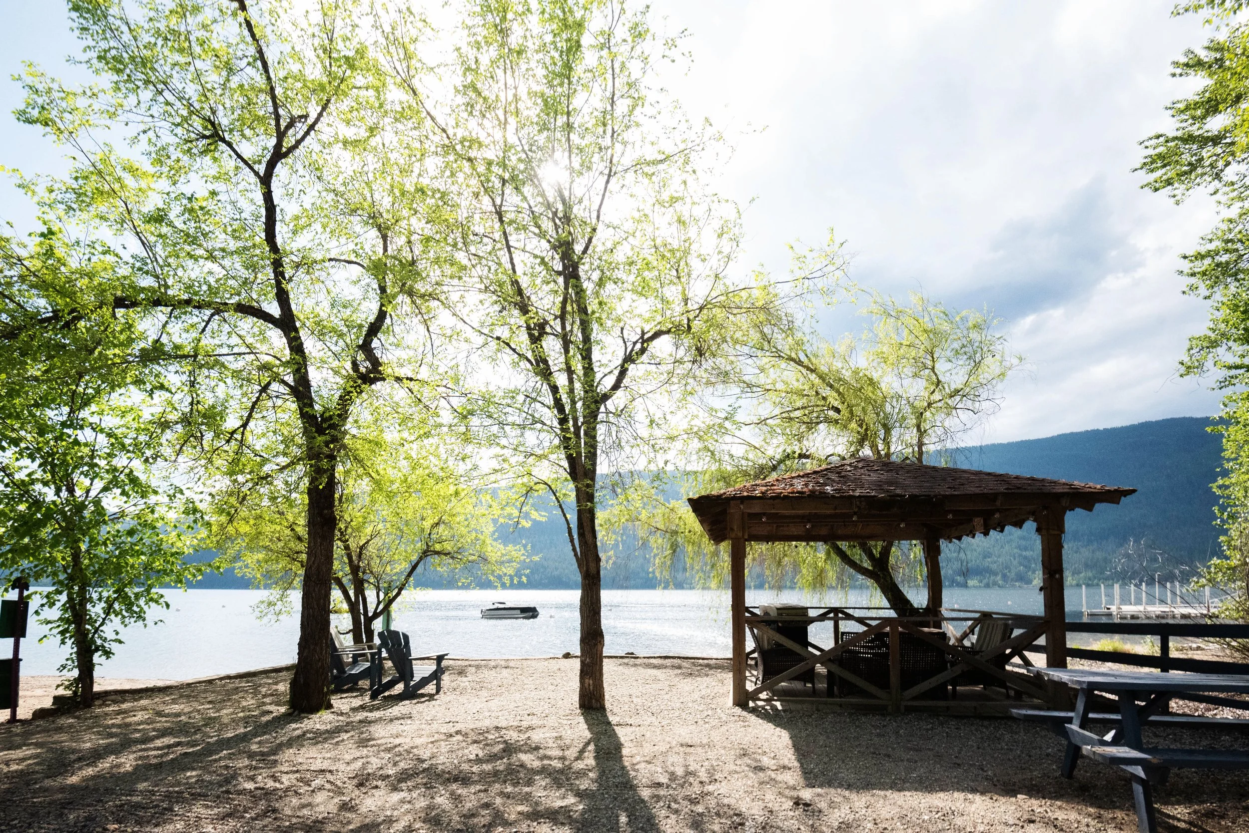 A lakeside scene with trees, a covered gazebo, chairs, dock, and boat in the water with mountains in the background.