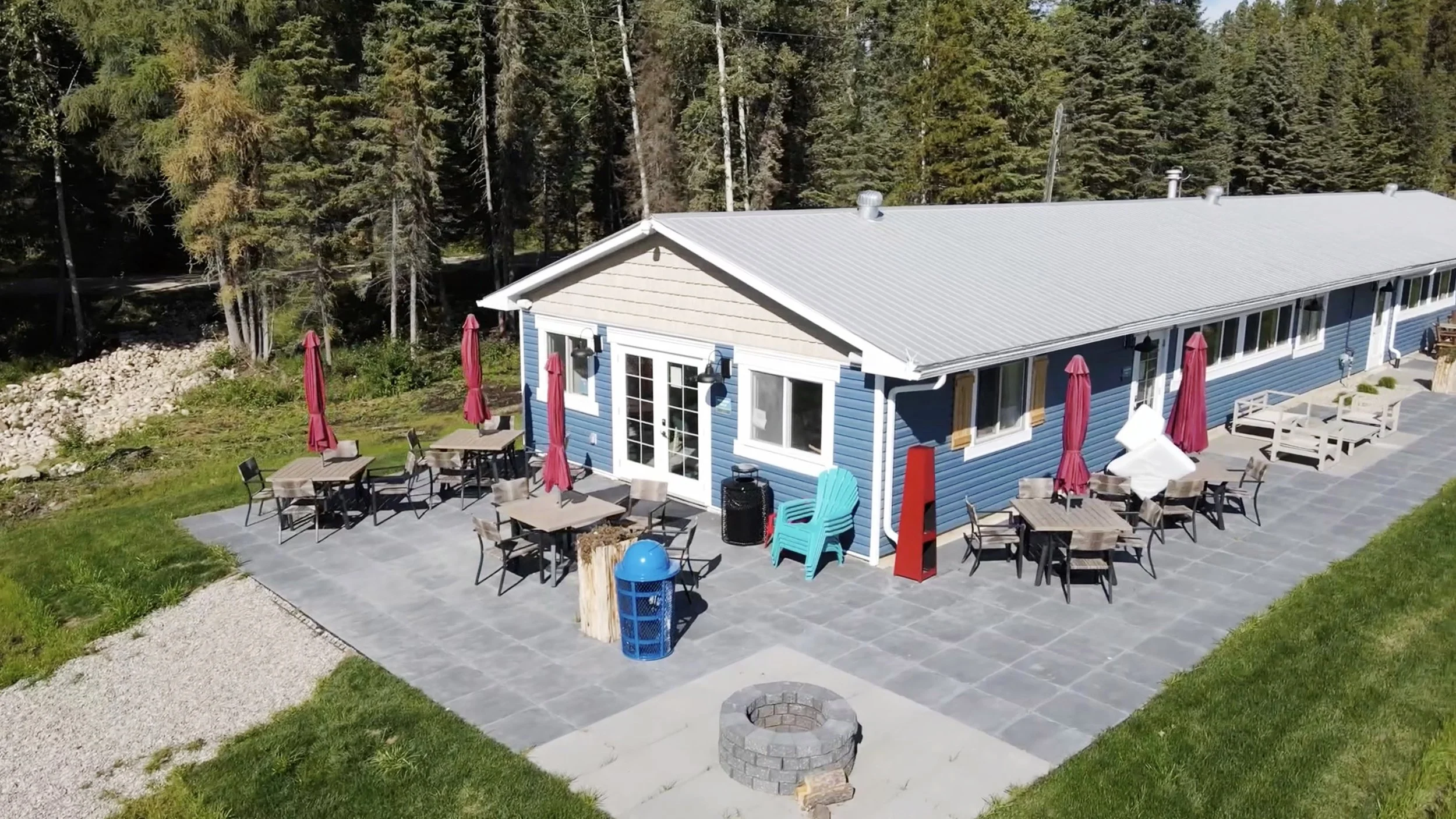 A blue house with a metal roof, outdoor patio with tables and chairs, red umbrellas, surrounded by a forested area with tall trees.