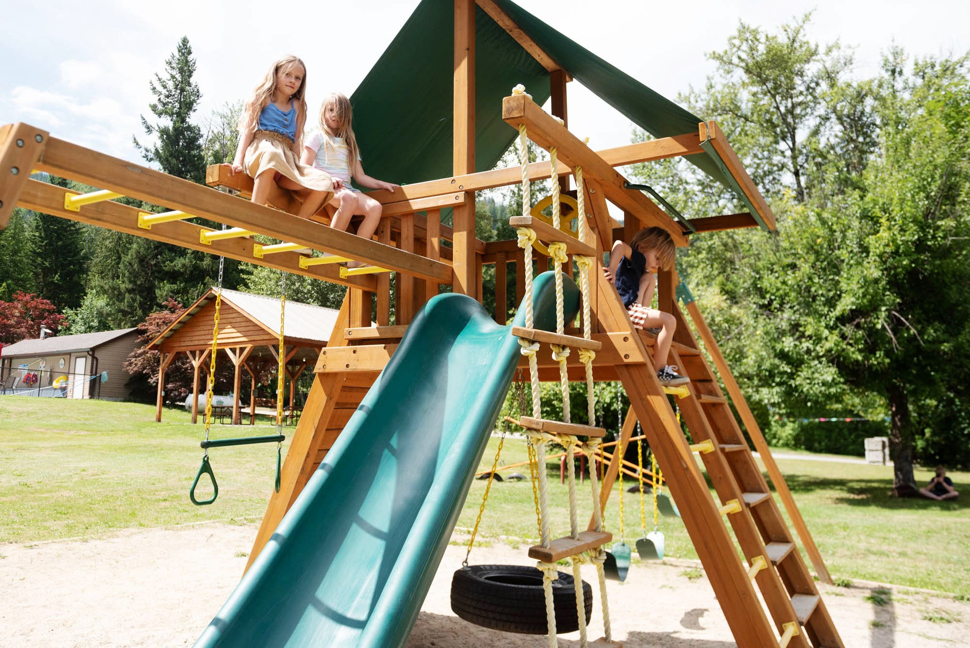 Children playing on a wooden playground structure with a slide, swings, and climbing elements, outdoors on a sunny day with trees in the background.