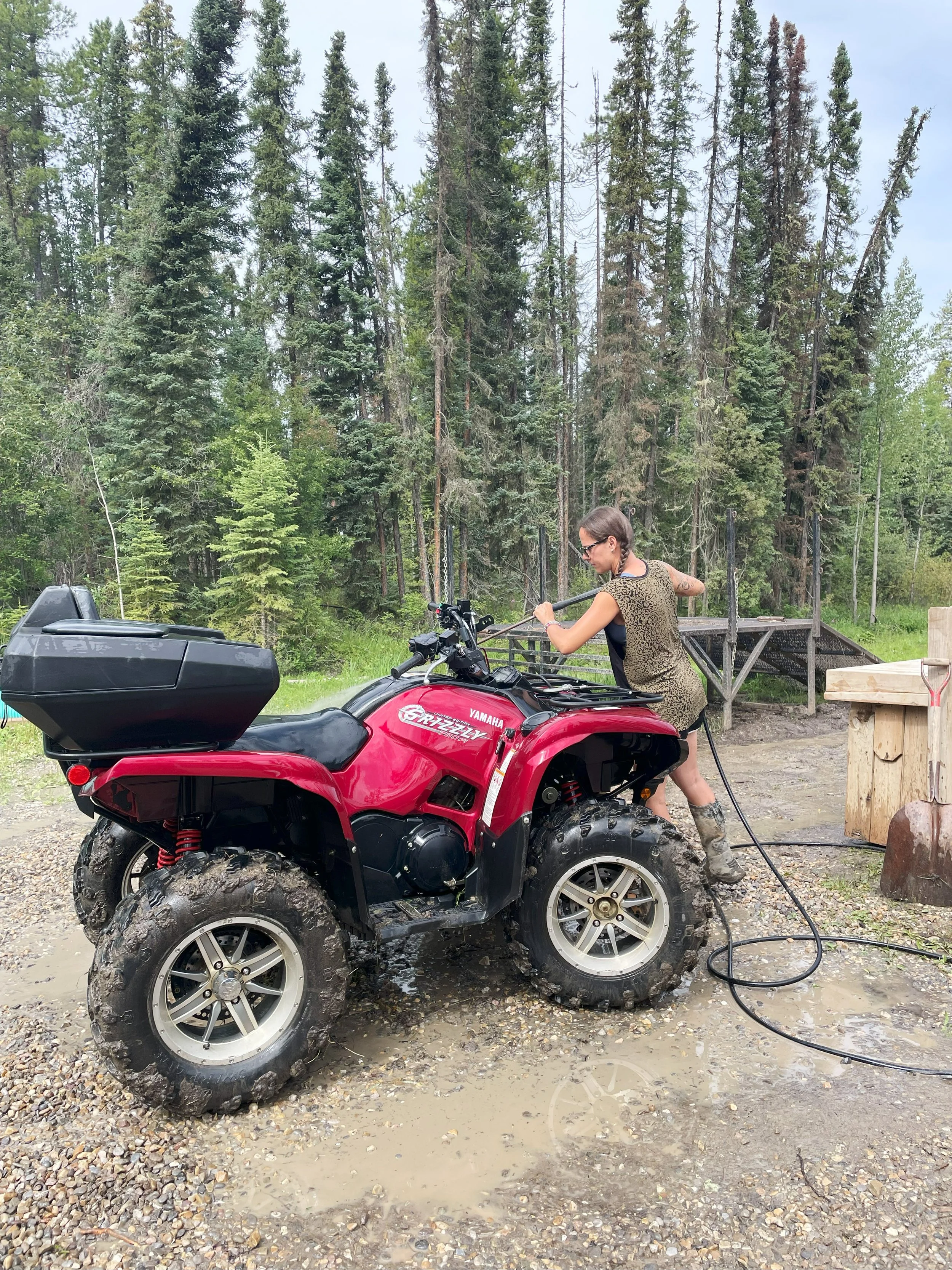 Woman cleaning a muddy red Yamaha Rhino 4-wheeler ATV outdoors amid trees, using a hose to wash it, with muddy water on the ground.