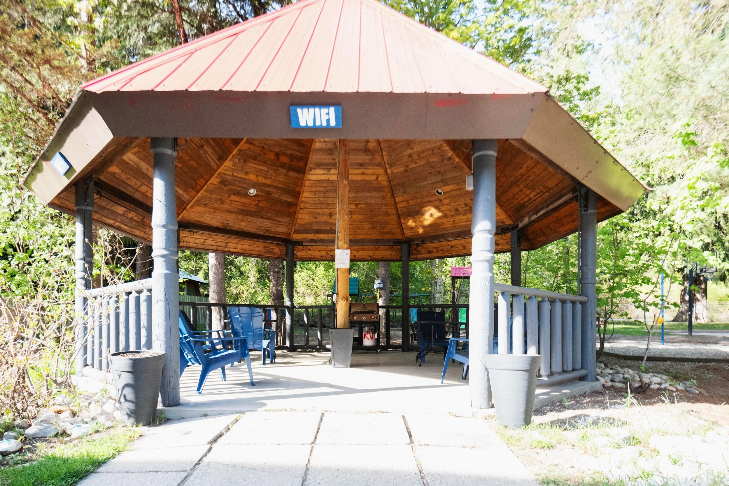 Outdoor pavilion with a red metal roof, supported by white columns, surrounded by trees and greenery, with blue chairs and a barbecue grill underneath.