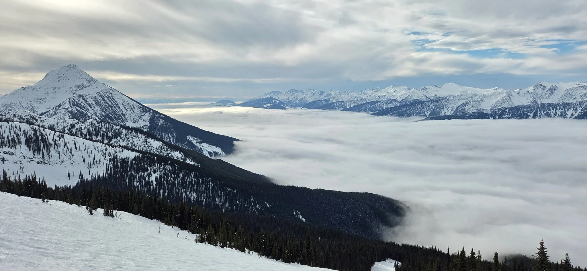 Snow-covered mountain range with clouds and fog in the valley below, evergreen trees on the slopes, and a partly cloudy sky.