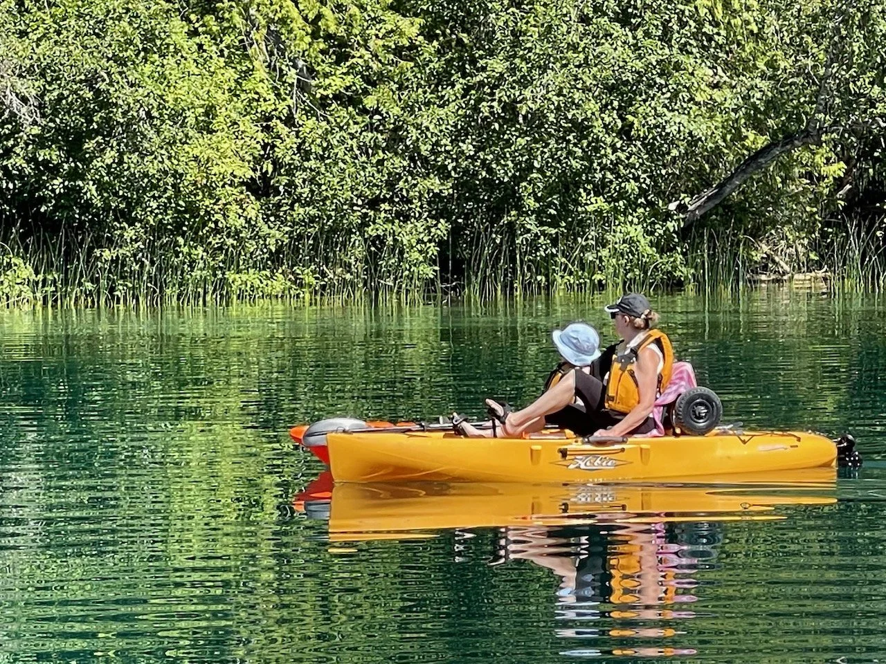 Two women kayaking on a calm body of water with dense trees and green foliage in the background.