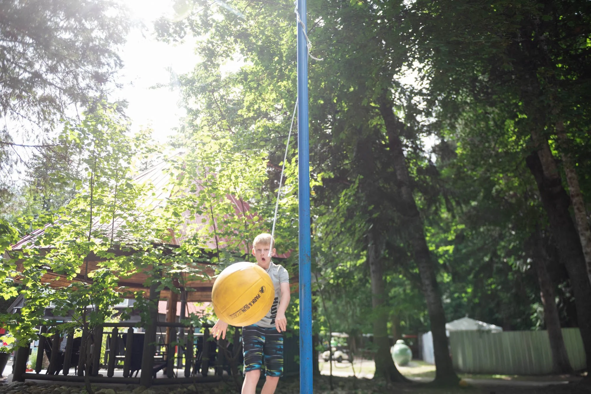 A boy playing volleyball outdoors, standing on grass with trees in the background, sunlight streaming through the leaves, holding a yellow volleyball.