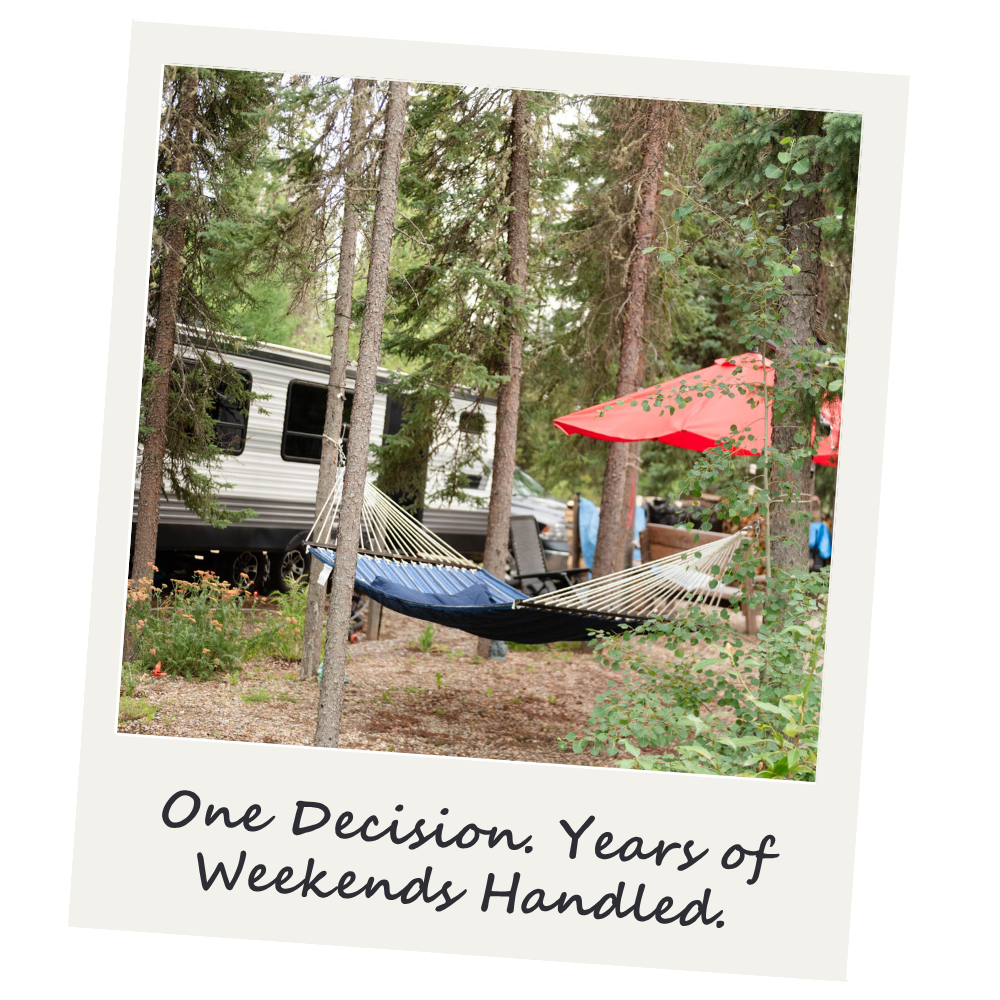 A hammock tied between two trees in a wooded area with a camper, orange umbrella, and camping chairs in the background.