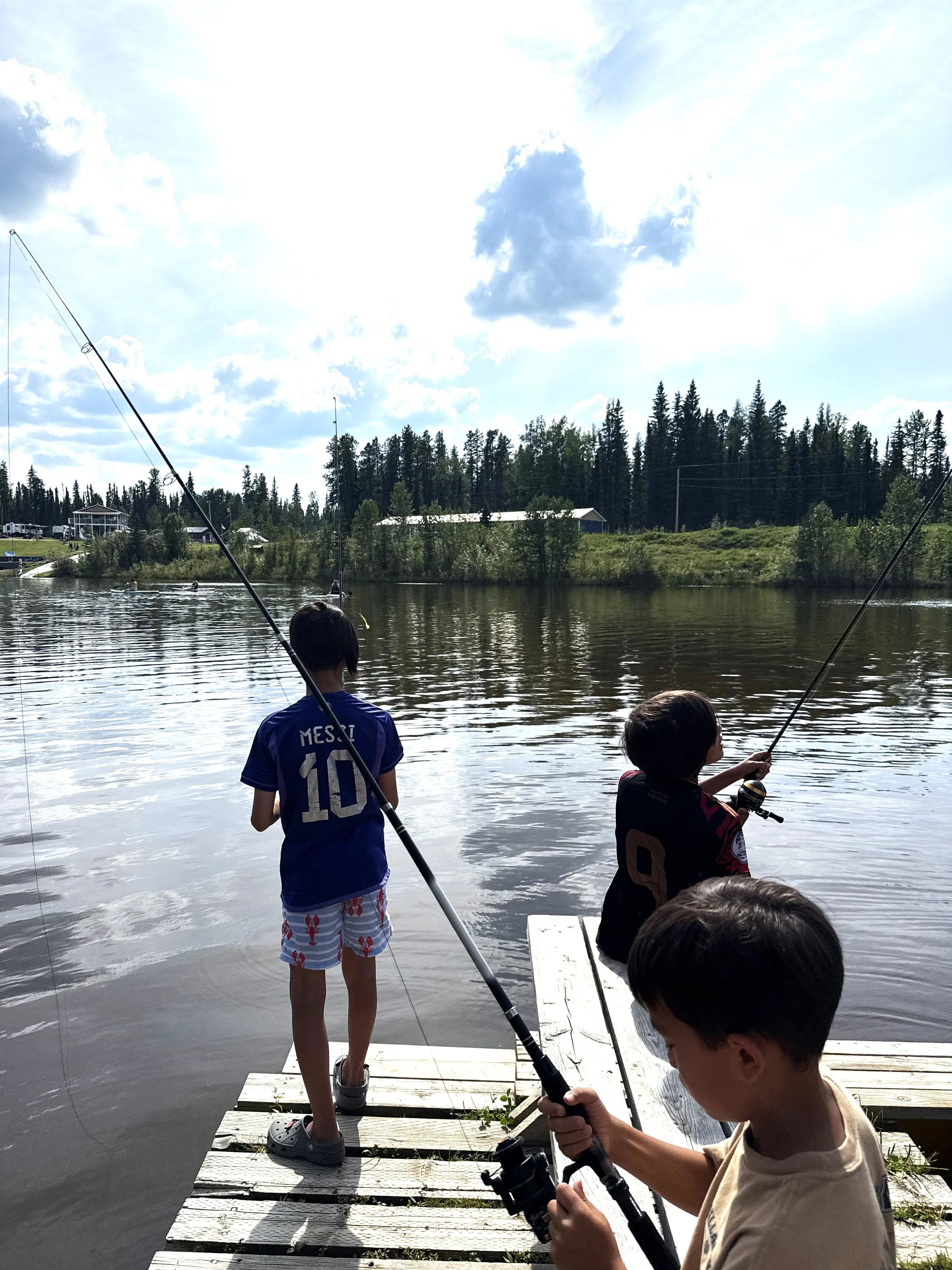 Children fishing off a dock by a lake, with trees and a building in the background under a partly cloudy sky.