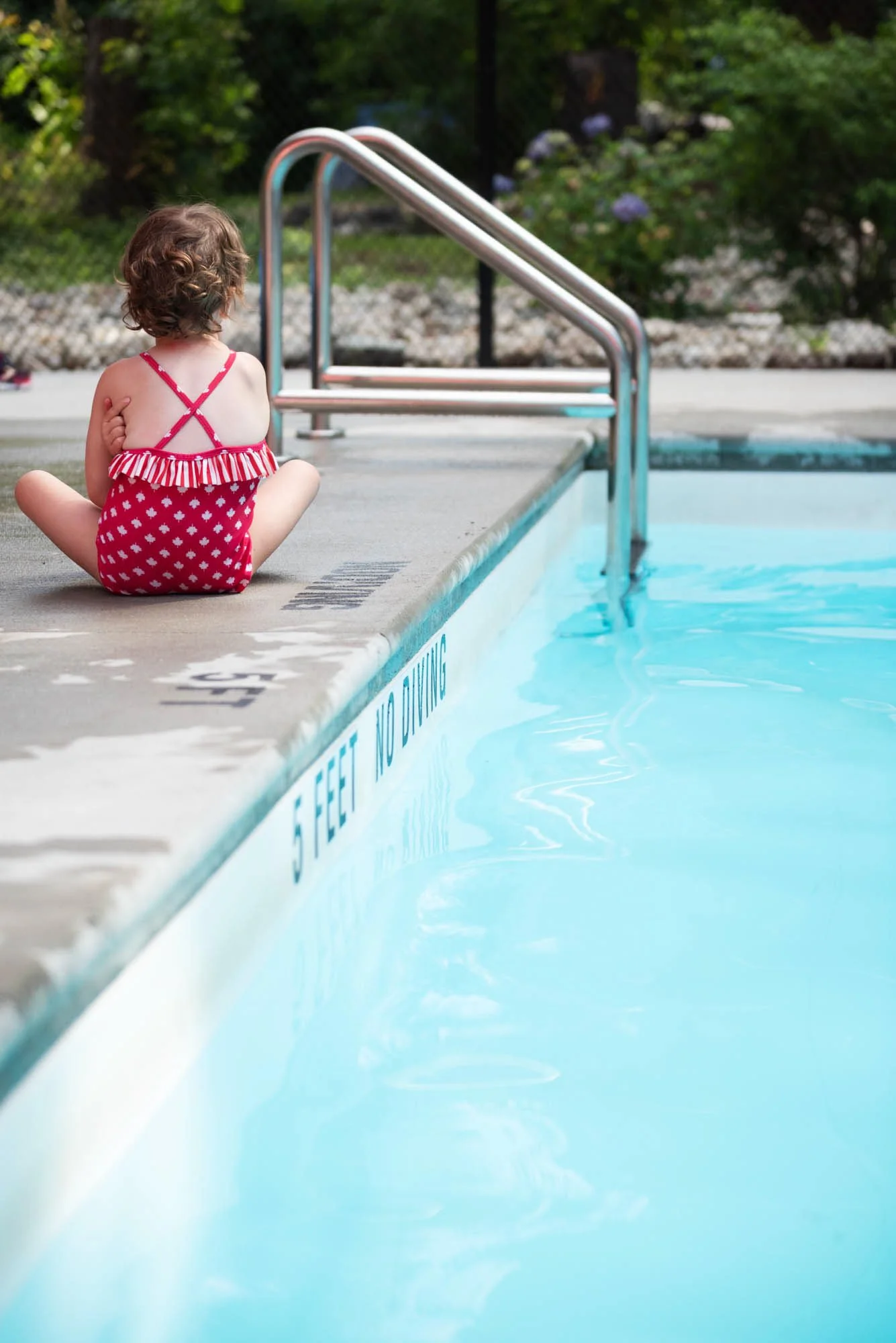 Young girl in red and white swimsuit sitting by a swimming pool, looking away, with green trees in the background.