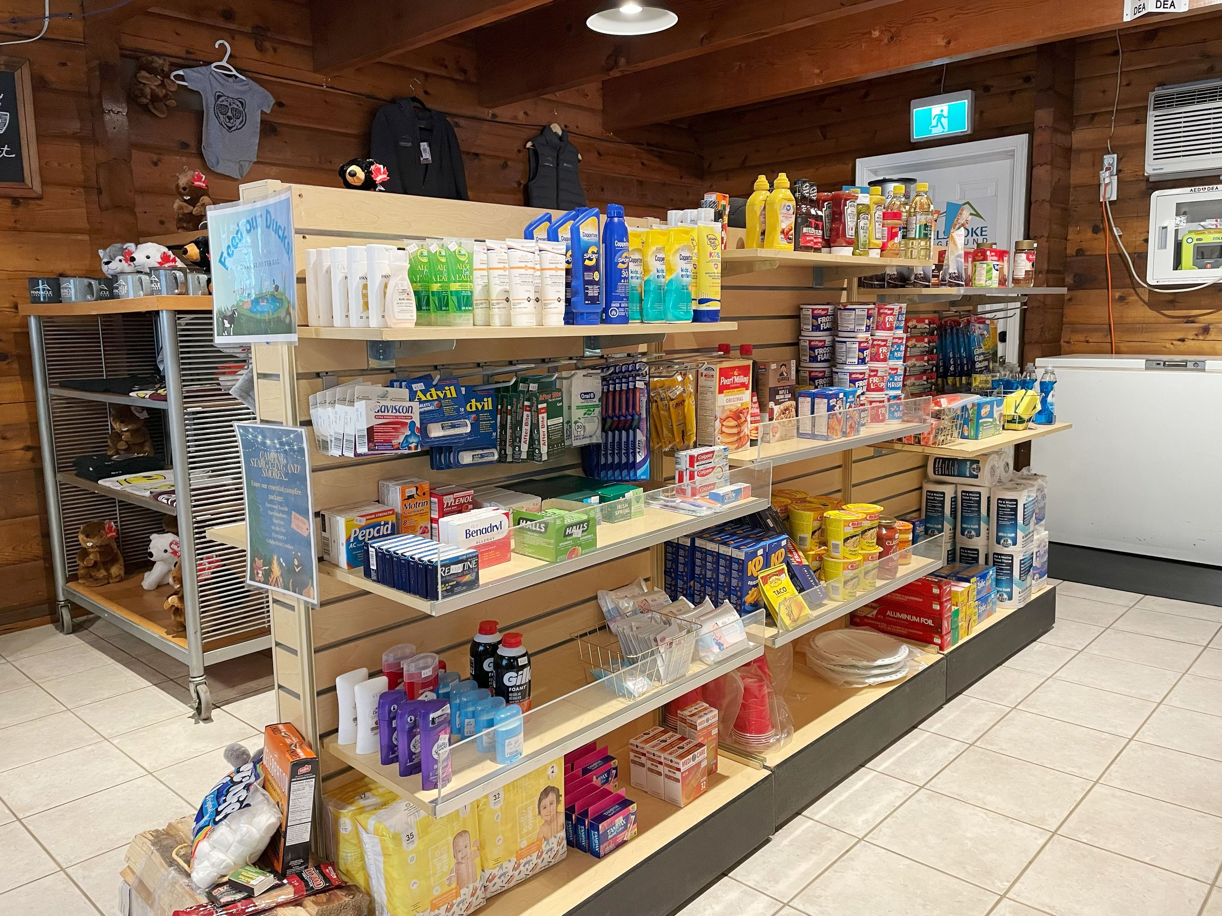 A grocery store aisle with shelves stocked with health and personal care products, snacks, and household items, against wood-paneled walls and tiled floors.