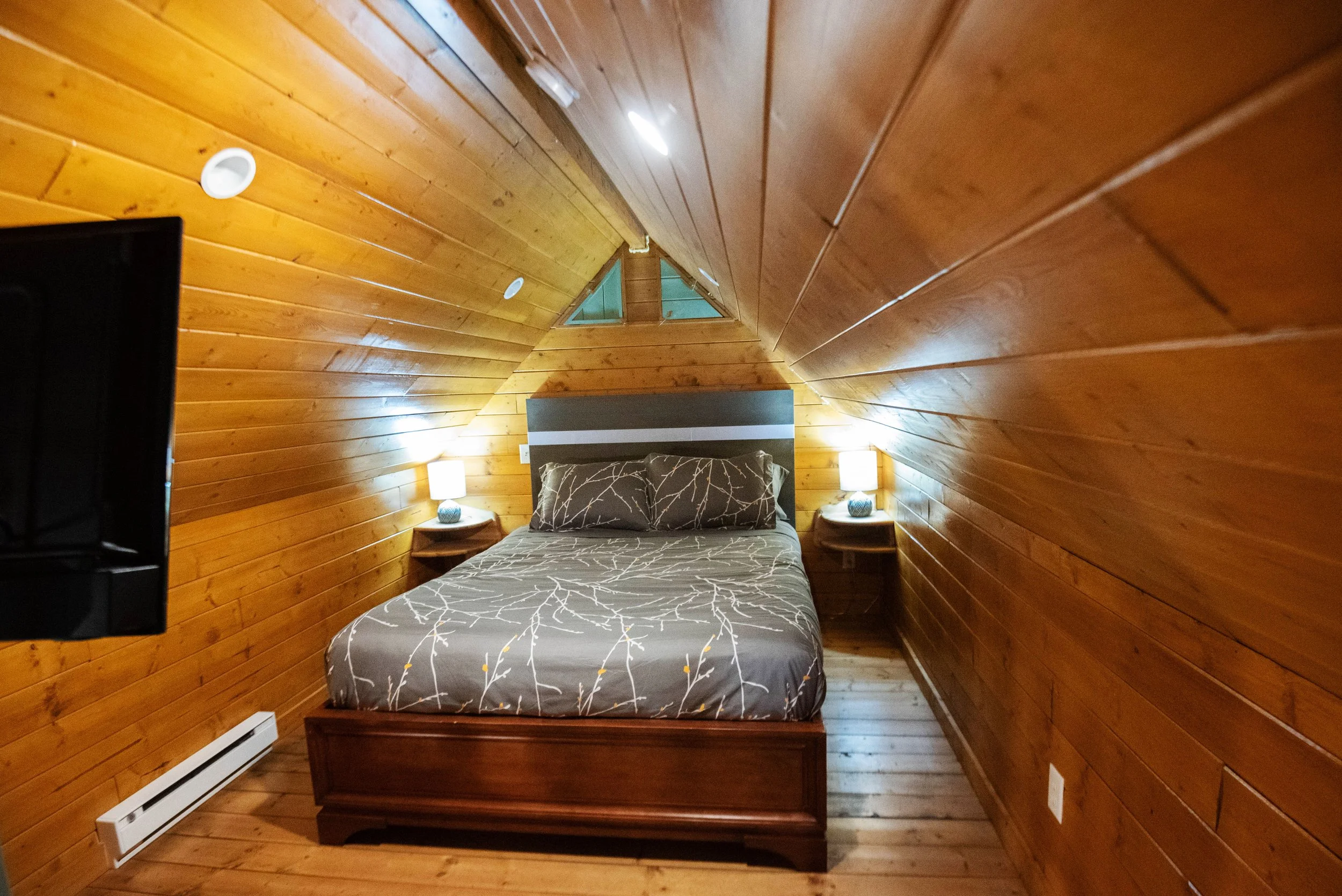 A cozy bedroom with wooden walls and ceiling, featuring a bed with patterned bedding, two nightstands with lamps, and a small triangular window above the headboard.