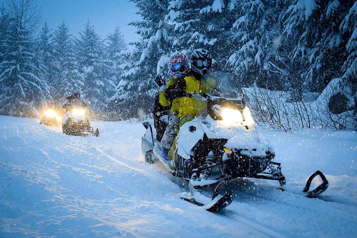 Two people riding snowmobiles through a snowy forest, with snow-covered trees and headlights illuminating the scene.