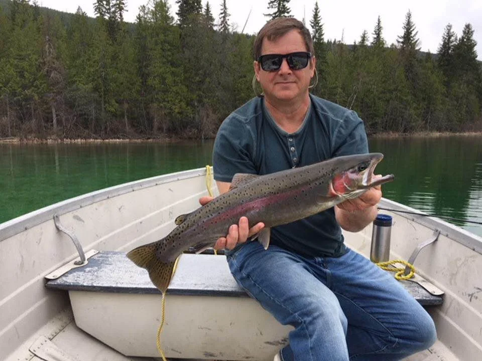 A man in sunglasses and a blue shirt sitting in a boat, holding a large rainbow trout fish with a scenic background of trees and water.