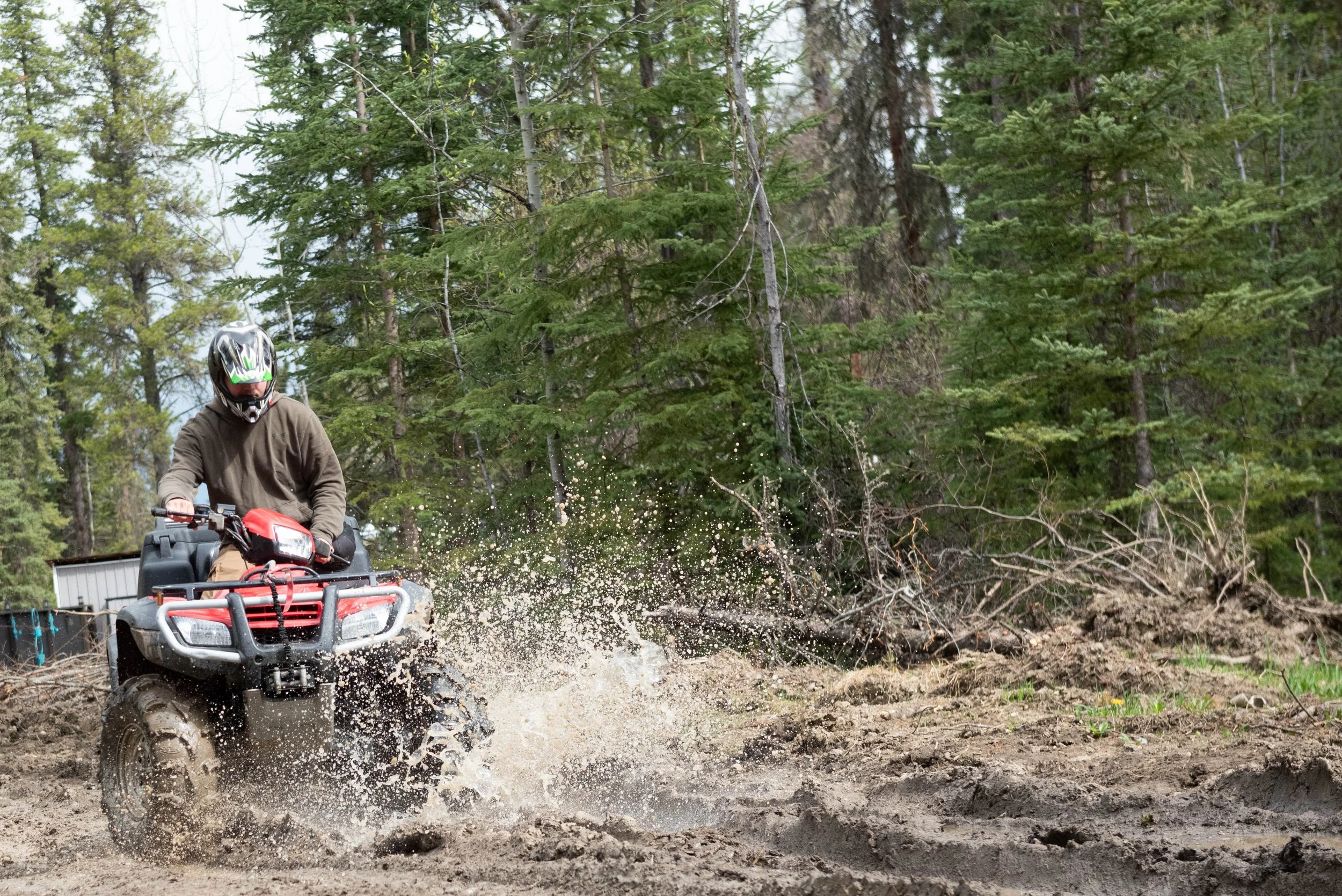 Person riding an all-terrain vehicle through a muddy outdoor trail surrounded by green trees, wearing a helmet and brown jacket.