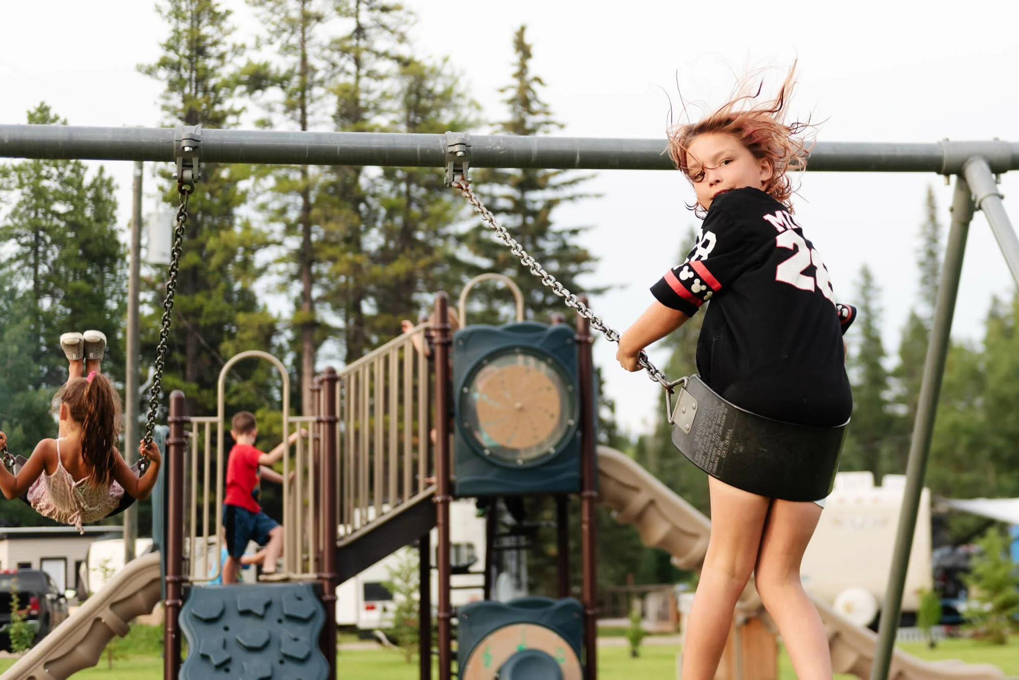 Kids playing on swings at a park, with a girl swinging and wind blowing through her hair, surrounded by trees and playground equipment.
