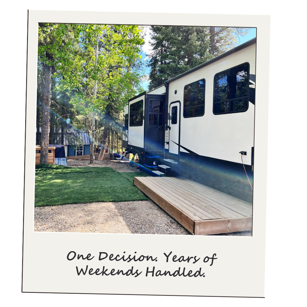 A travel trailer parked on a dirt and grass area in a wooded setting with tall trees and blue sky. The trailer has a small wooden deck in front of the door. The caption reads, "One Decision. Years of Weekends Handled."