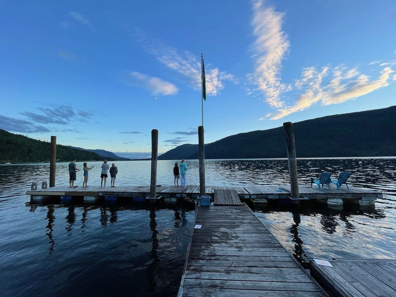 People standing and taking photos on a dock by a body of water, with mountains in the background and a partly cloudy sky overhead.