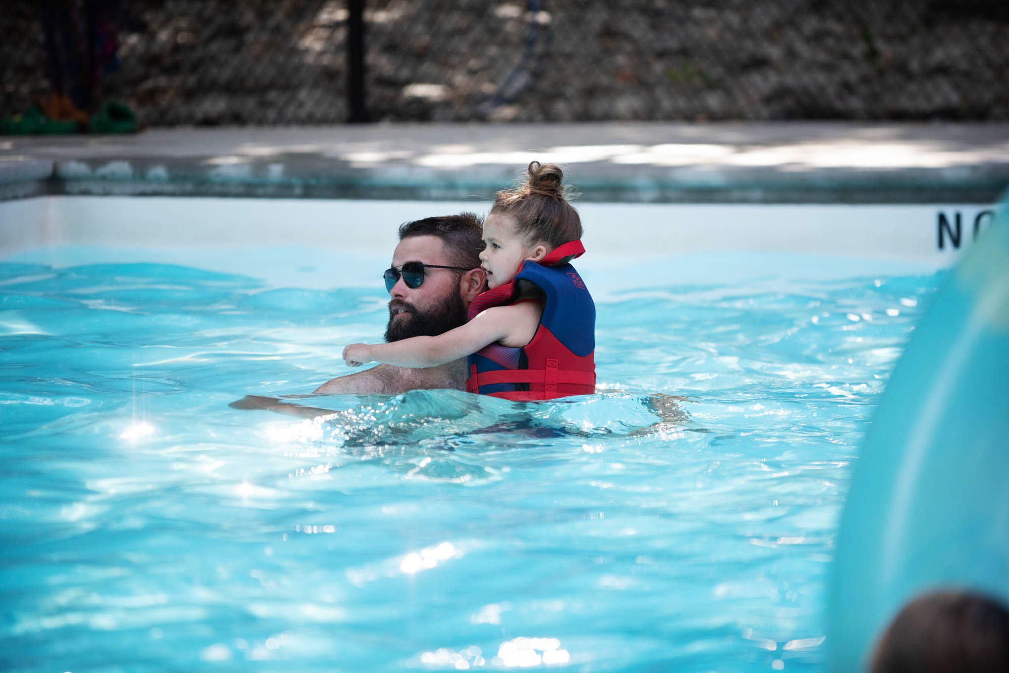 A man with sunglasses is in a swimming pool holding a young girl with a top bun hairstyle, who is wearing a life jacket.