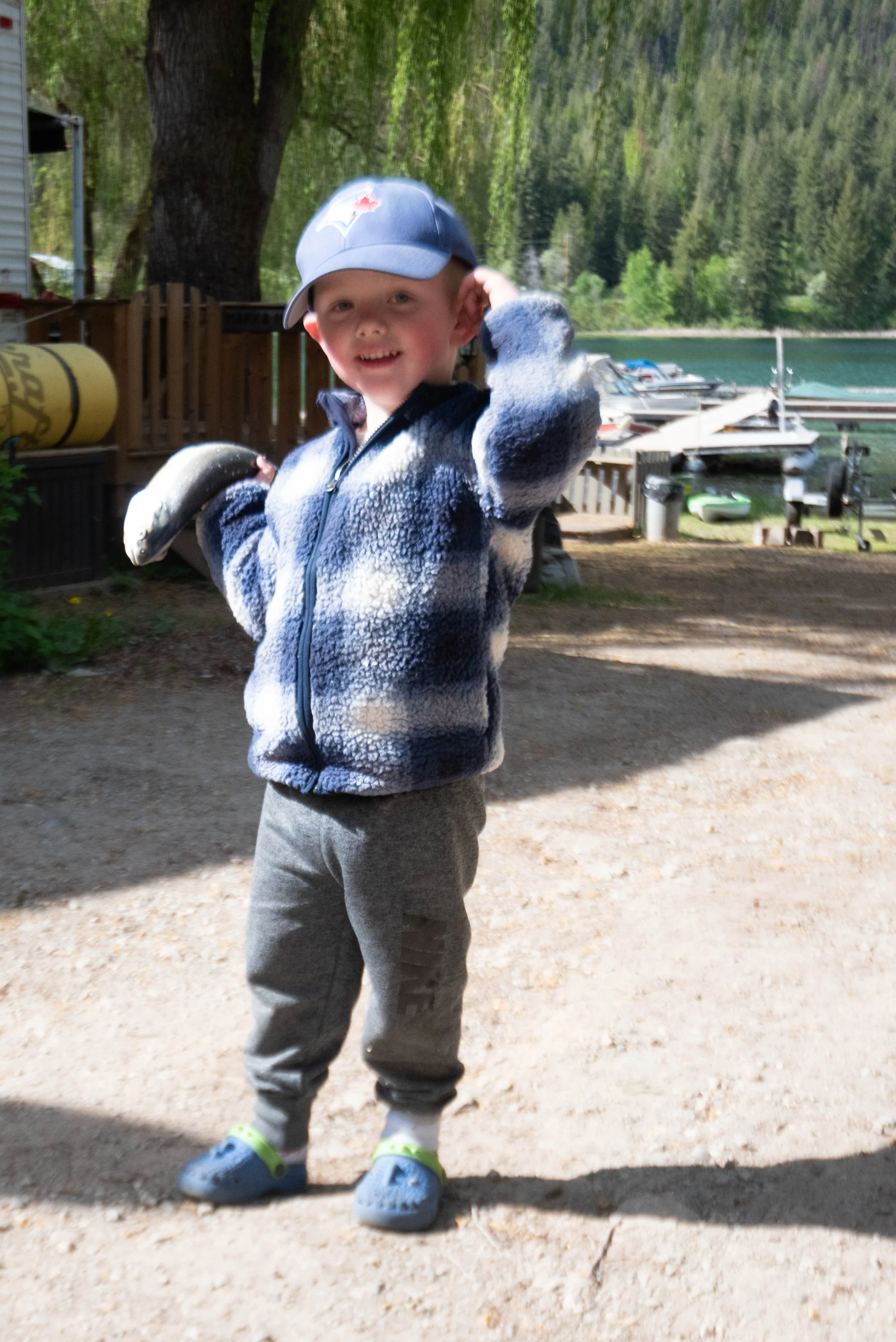 A young boy wearing a blue baseball cap, a fleece jacket, gray pants, and Crocs shoes, standing outdoors near a lake with boats and a forest in the background.