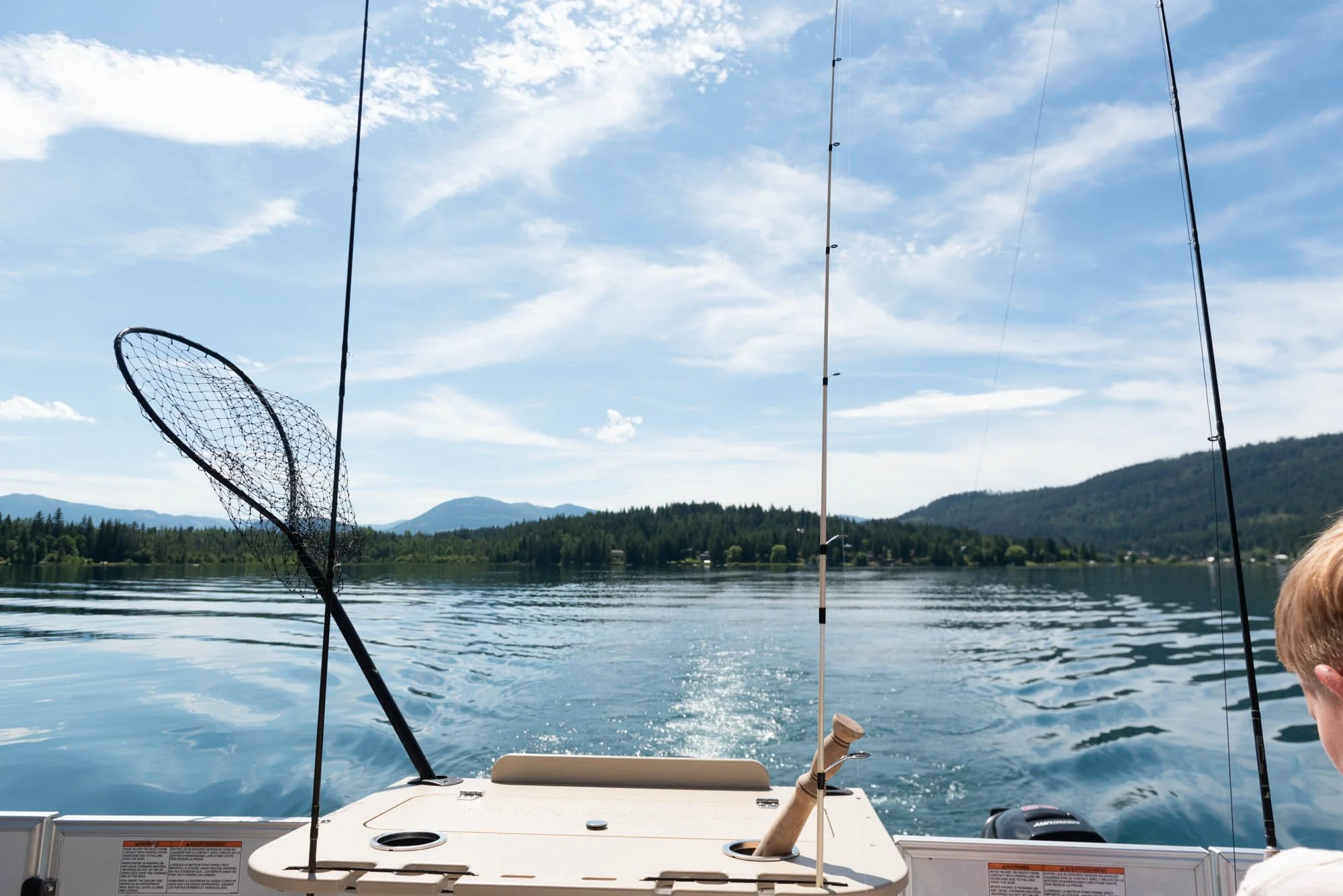A fishing boat on a lake with fishing rods and a net, with a backdrop of mountains and trees under a partly cloudy sky.