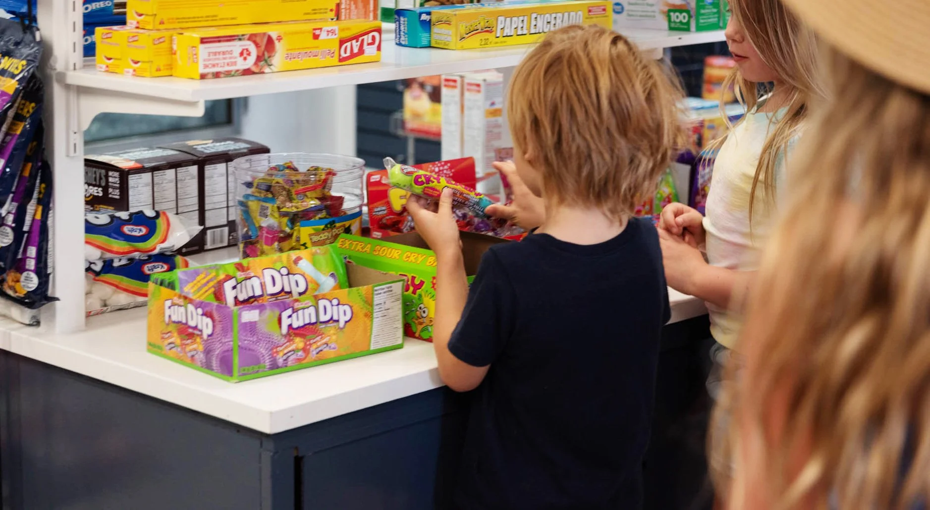 Two children, a girl and a boy, at a candy counter in a store, choosing colorful candy packages among shelves of snacks and paper products.