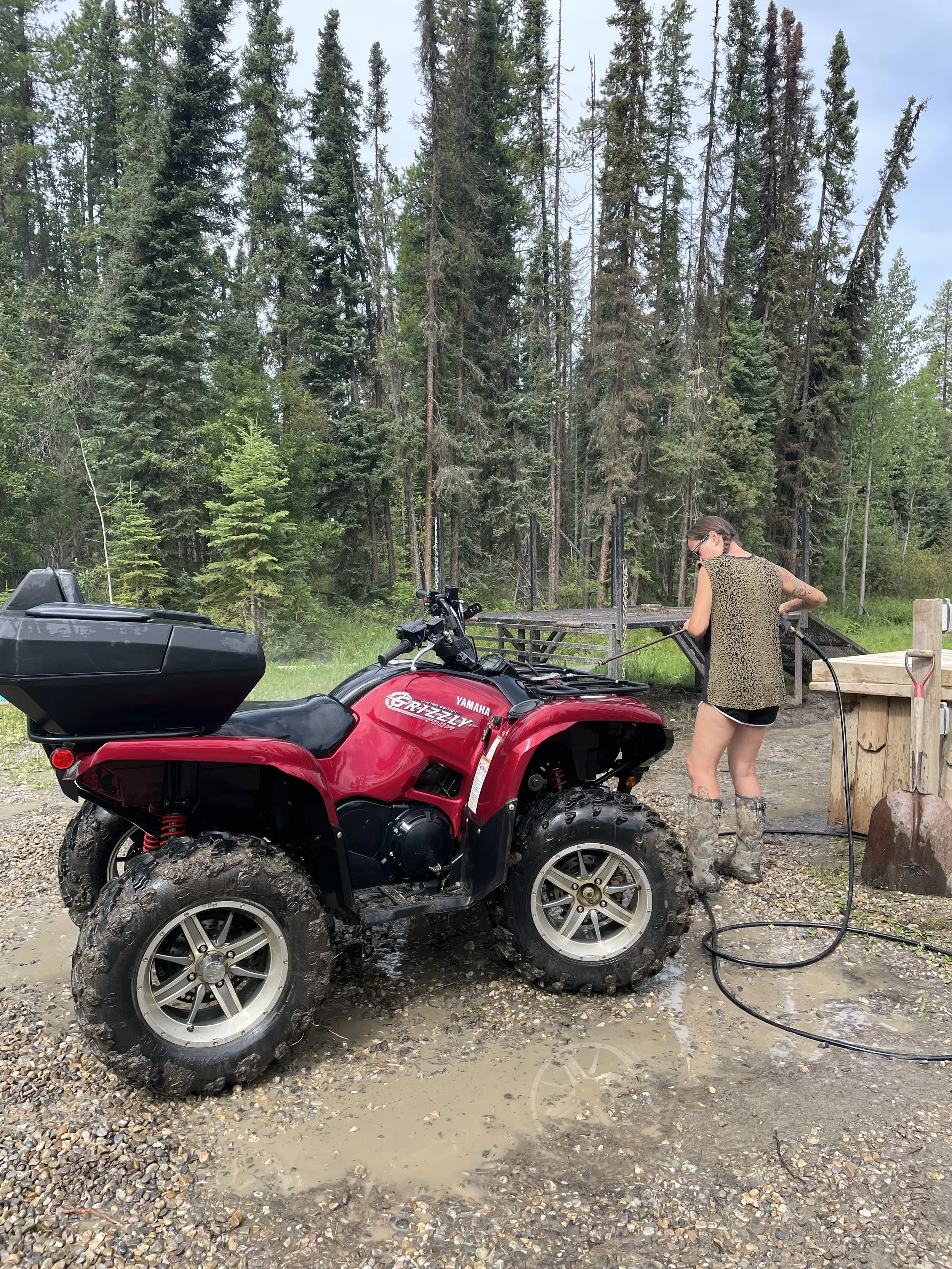 A woman filling an ATV with water at an outdoor location surrounded by trees. The ATV has muddy tires and a red body, with a black storage box on the back. The woman is wearing a sleeveless leopard print top, black shorts, and rubber boots.