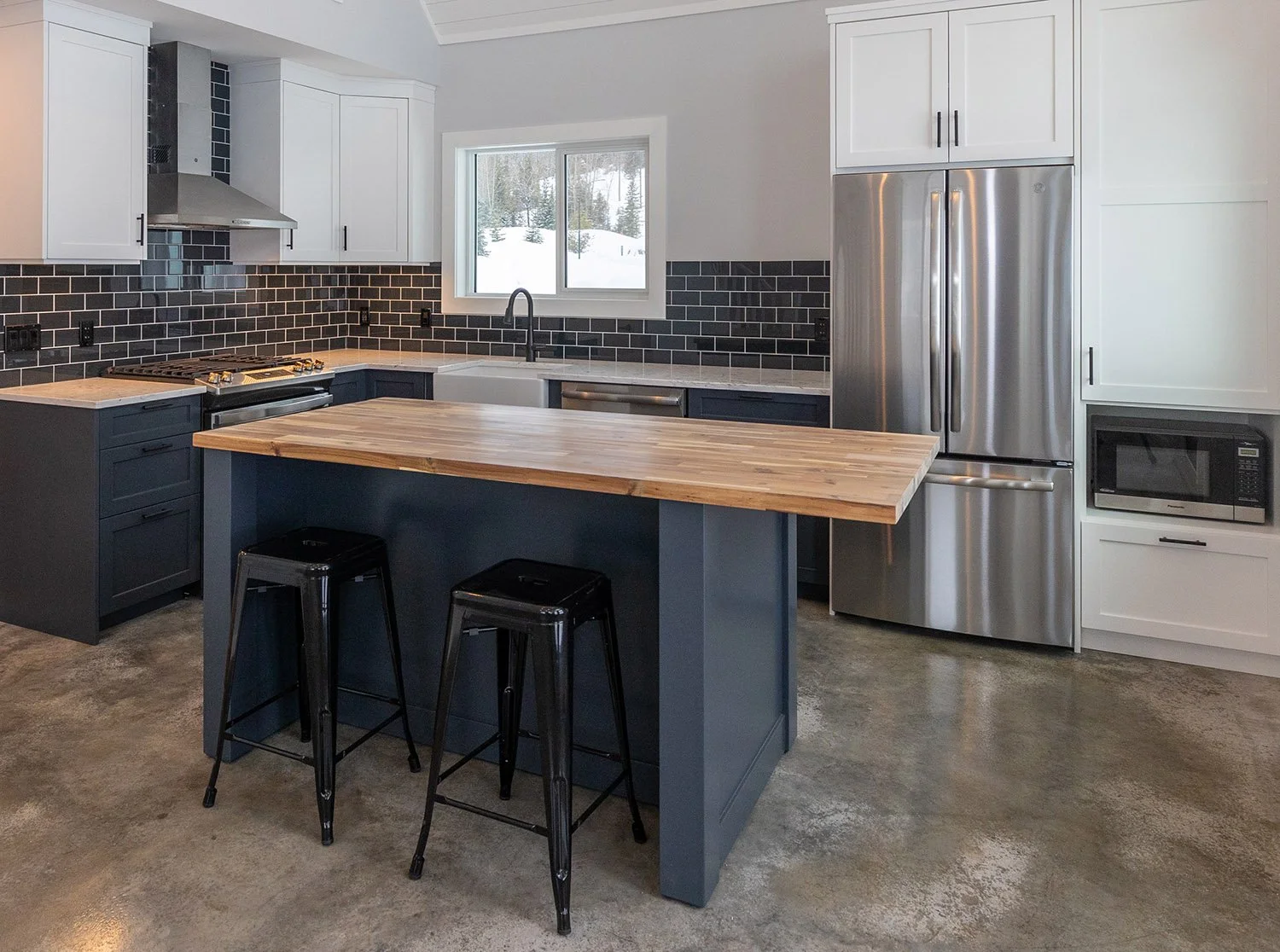 Modern kitchen with white upper cabinets, dark lower cabinets, black subway tile backsplash, stainless steel refrigerator, microwave, and range hood, kitchen island with a wooden countertop, two black barstools, window above sink showing snowy outdoo