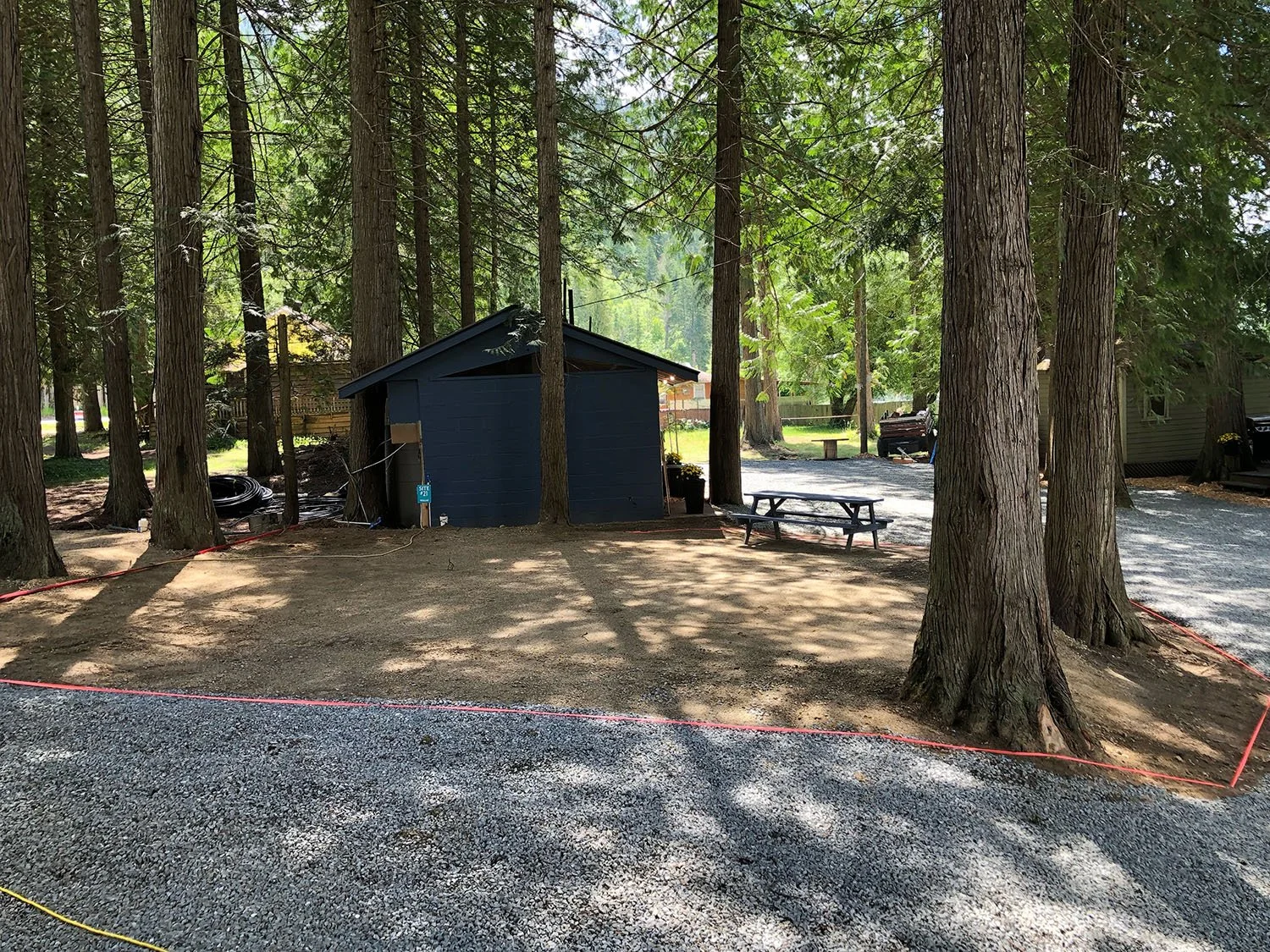A shaded outdoor area with tall trees, a small blue shed, picnic tables, and gravel ground, with some construction markers on the ground.