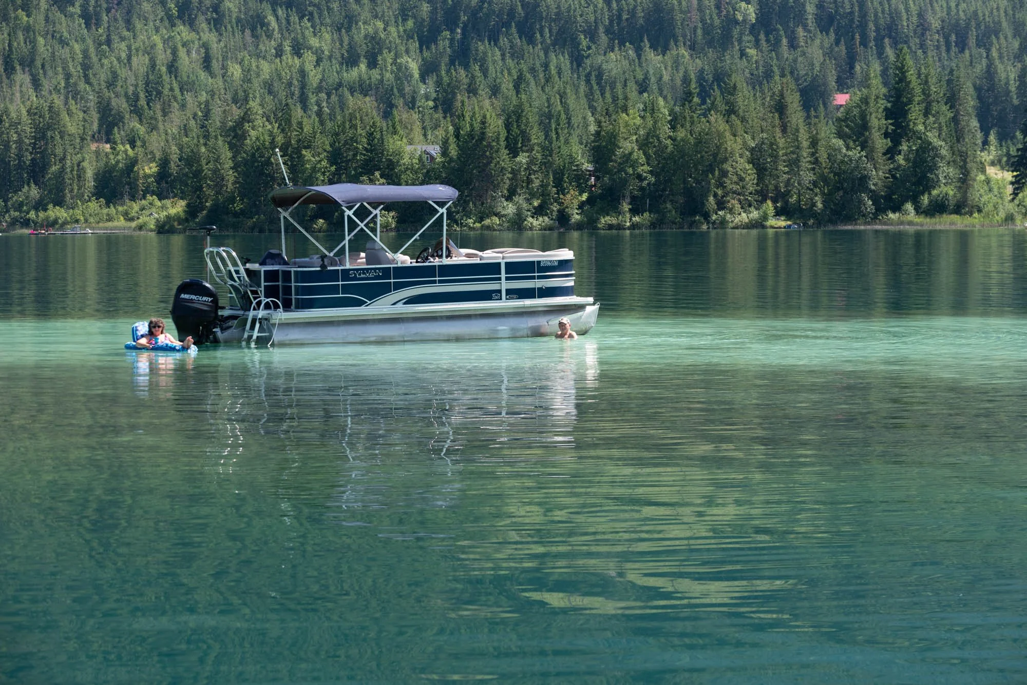A blue pontoon boat named 'Sylvan' floating on a calm green lake with two people swimming nearby and a forested hillside in the background.