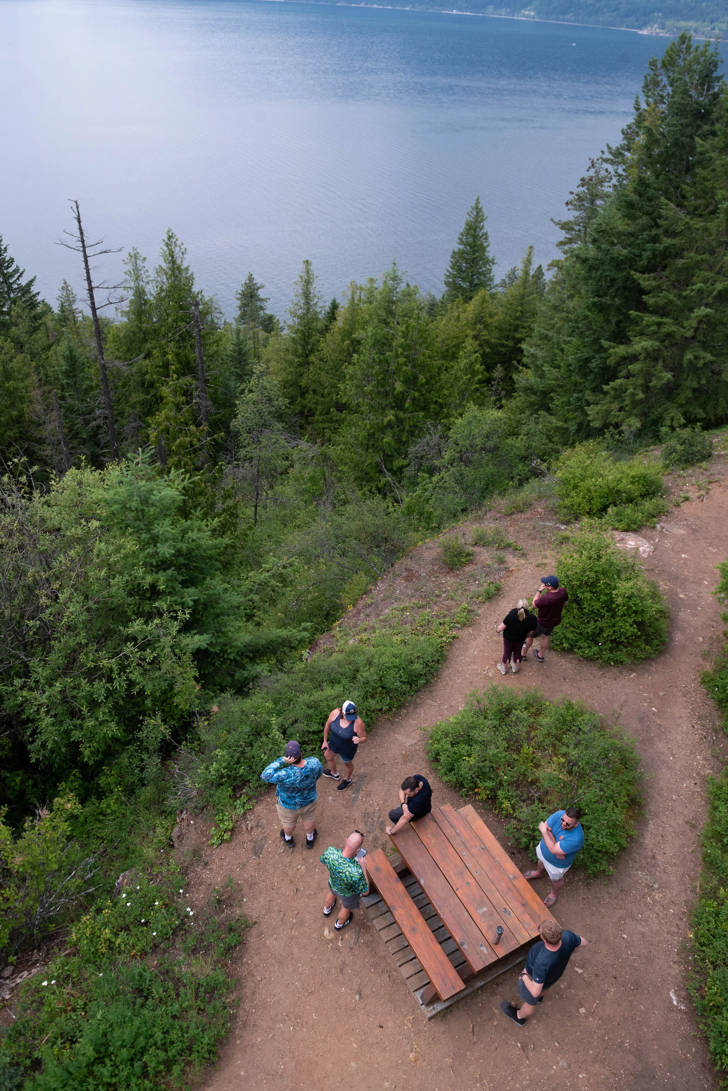Bird's-eye view of a group of people standing and talking on a dirt trail near a lake, surrounded by dense green trees and hills in the background.