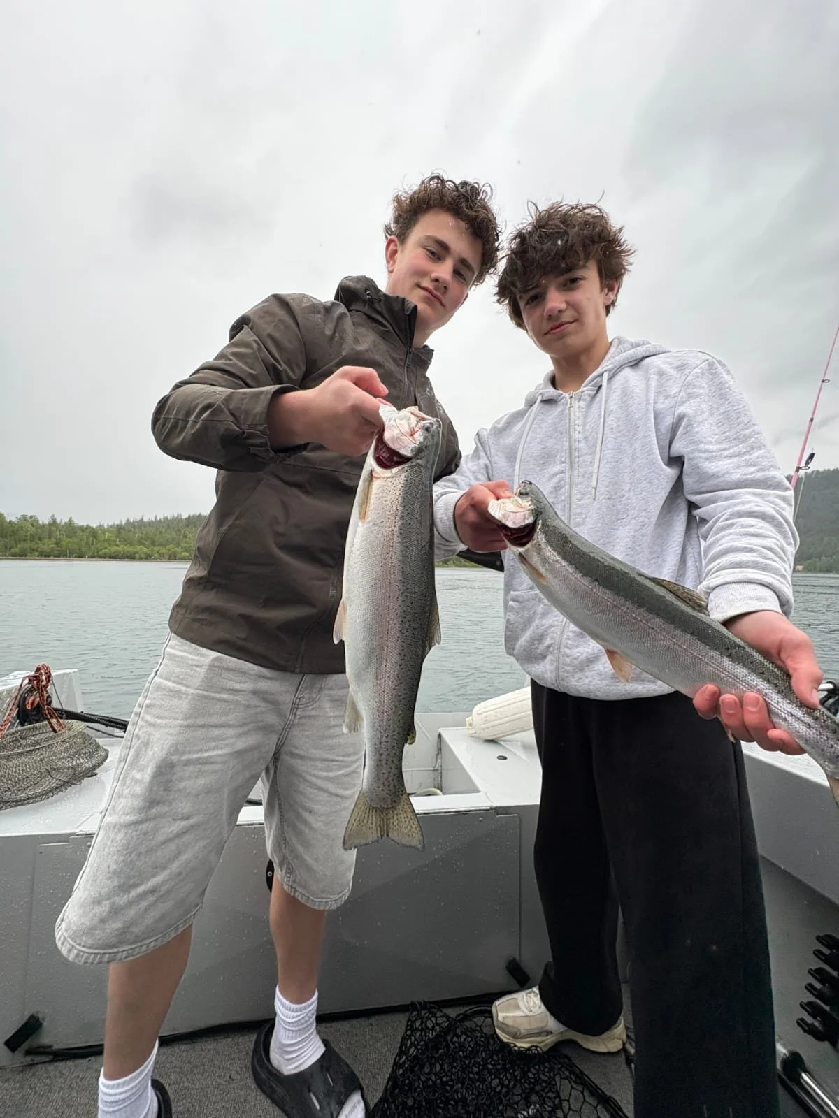 Two young men on a boat holding large fish, smiling and displaying their catch during a cloudy day. One wears a brown jacket, the other a gray hoodie.