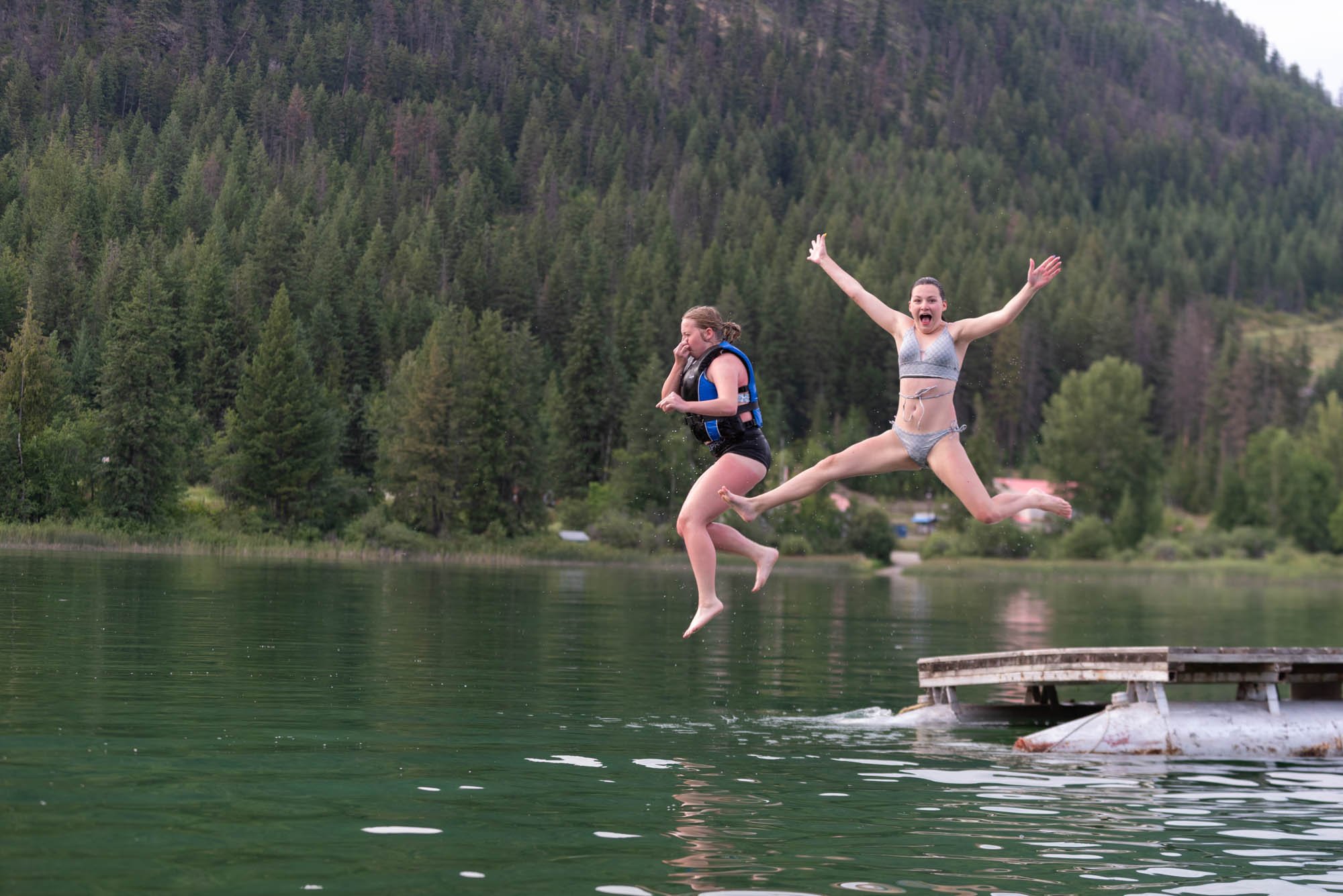 Two girls jumping off a dock into a lake with a background of forested hills.