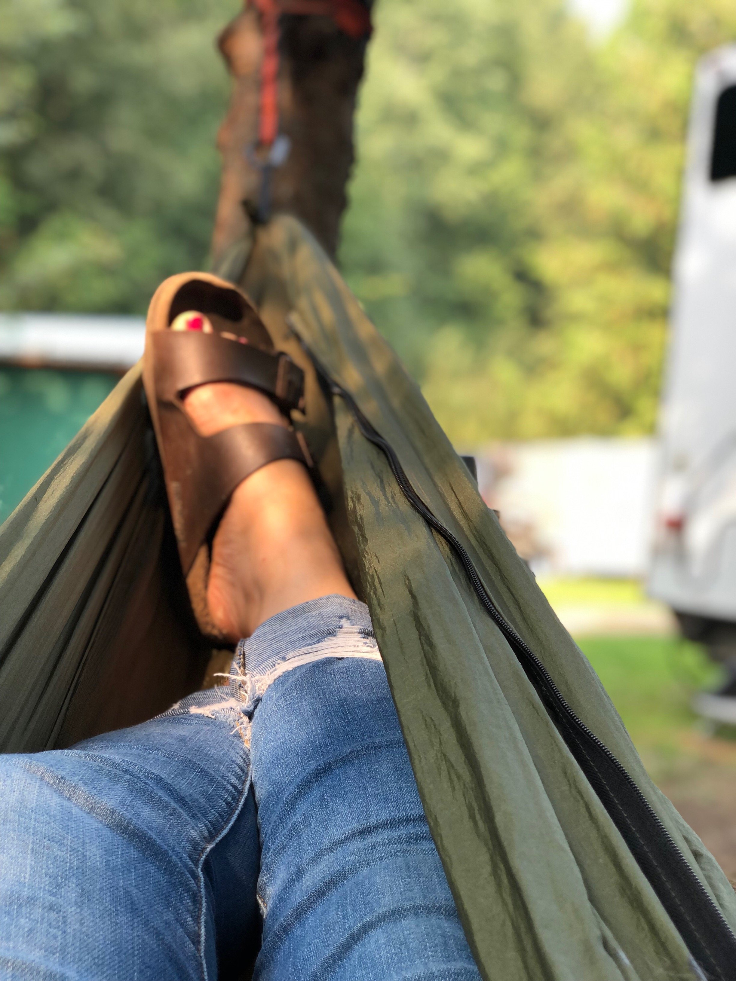Person lying in a hammock outdoors, wearing denim jeans and brown sandals, with green trees and a parked vehicle in the background.