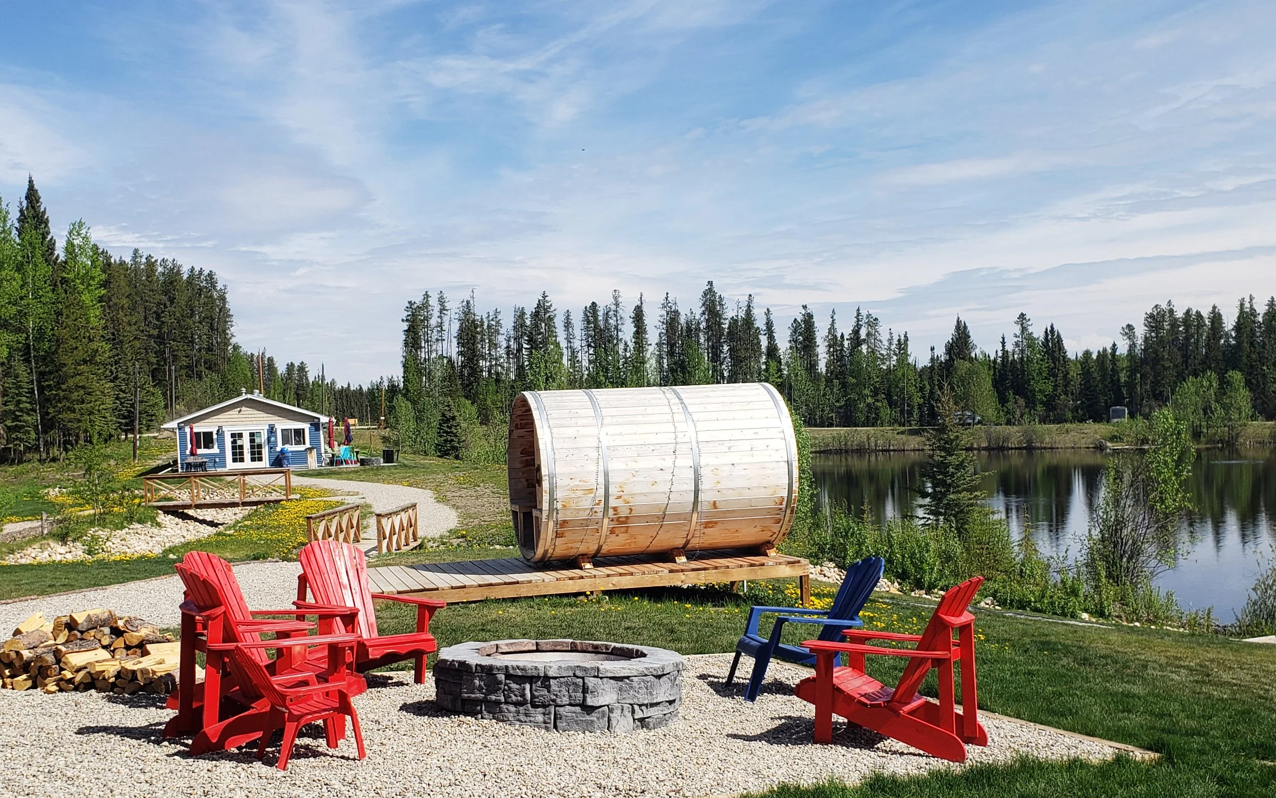 Outdoor scene with red Adirondack chairs around a fire pit, wood fire bundle nearby, a wooden sauna barrel, a small house with a deck and outdoor furniture, a pond with trees on the opposite bank, and a clear blue sky with some clouds.