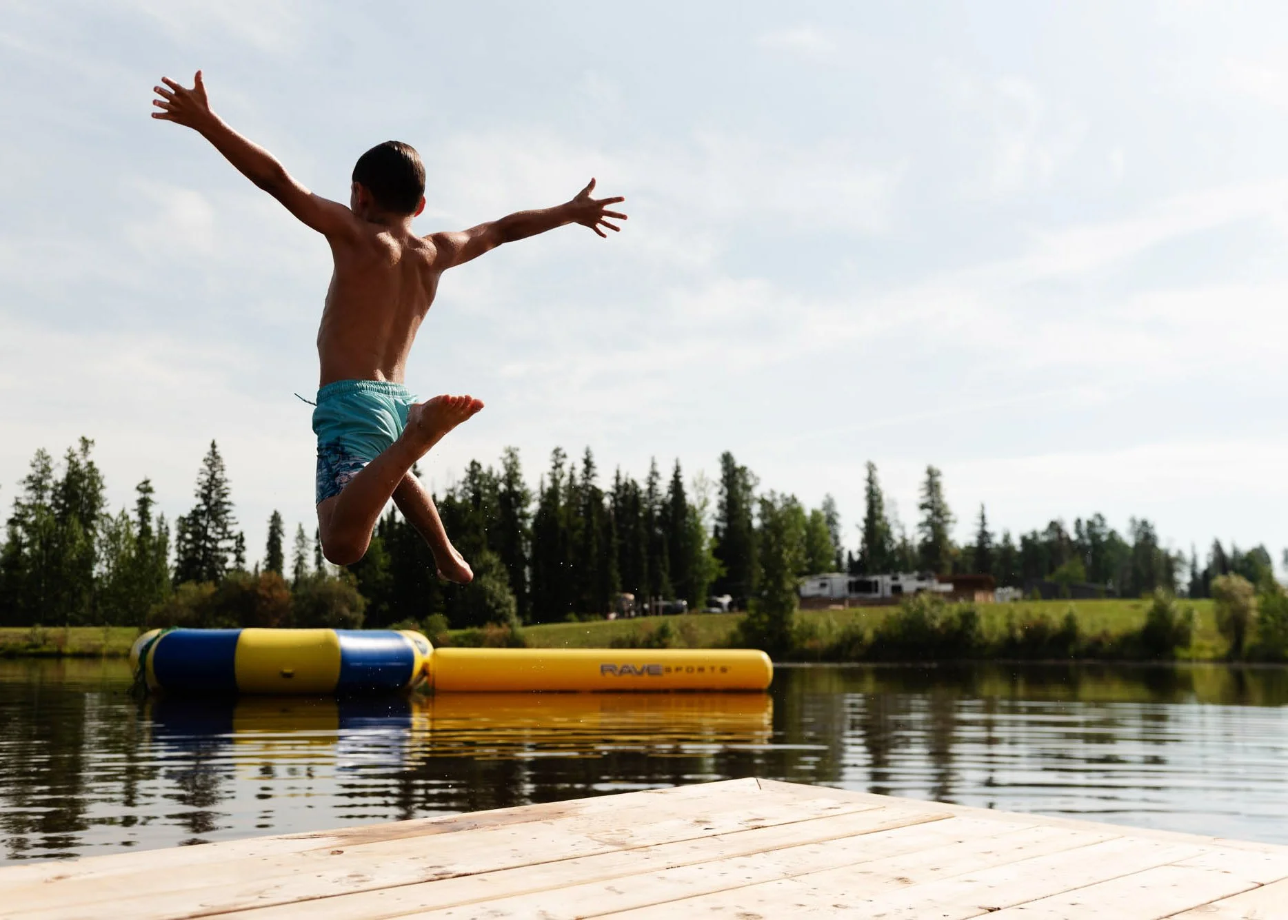 A young boy in swim trunks jumping into a lake from a wooden dock, with trees and houses in the background on a cloudy day.