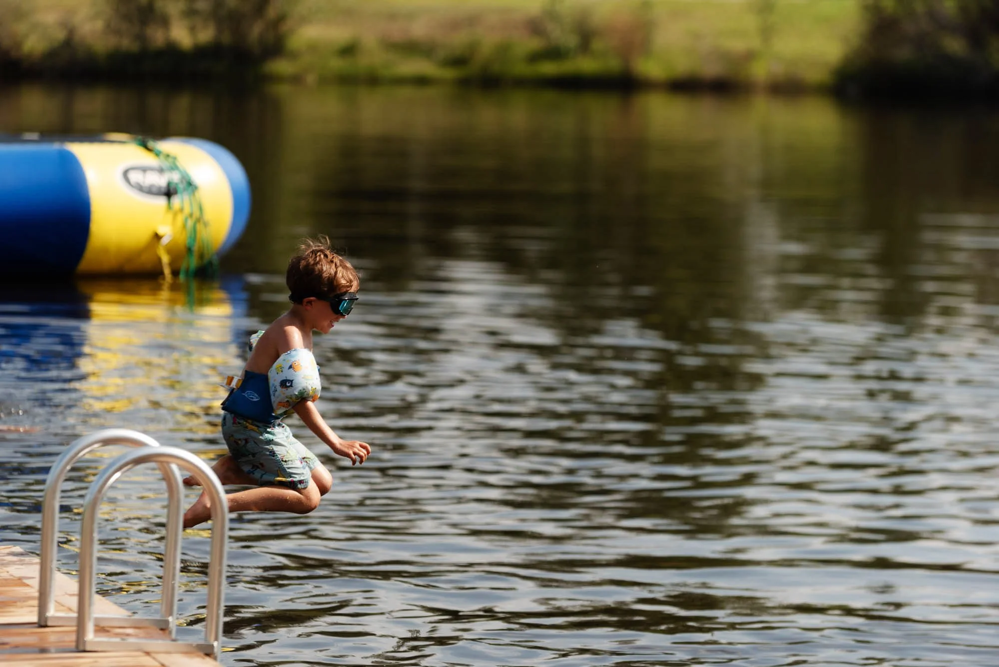 A young boy sitting on the edge of a dock by a lake, wearing swim trunks and goggles, with one foot in the water. In the background, there is a yellow and blue inflatable float and trees on the far shore of the lake.